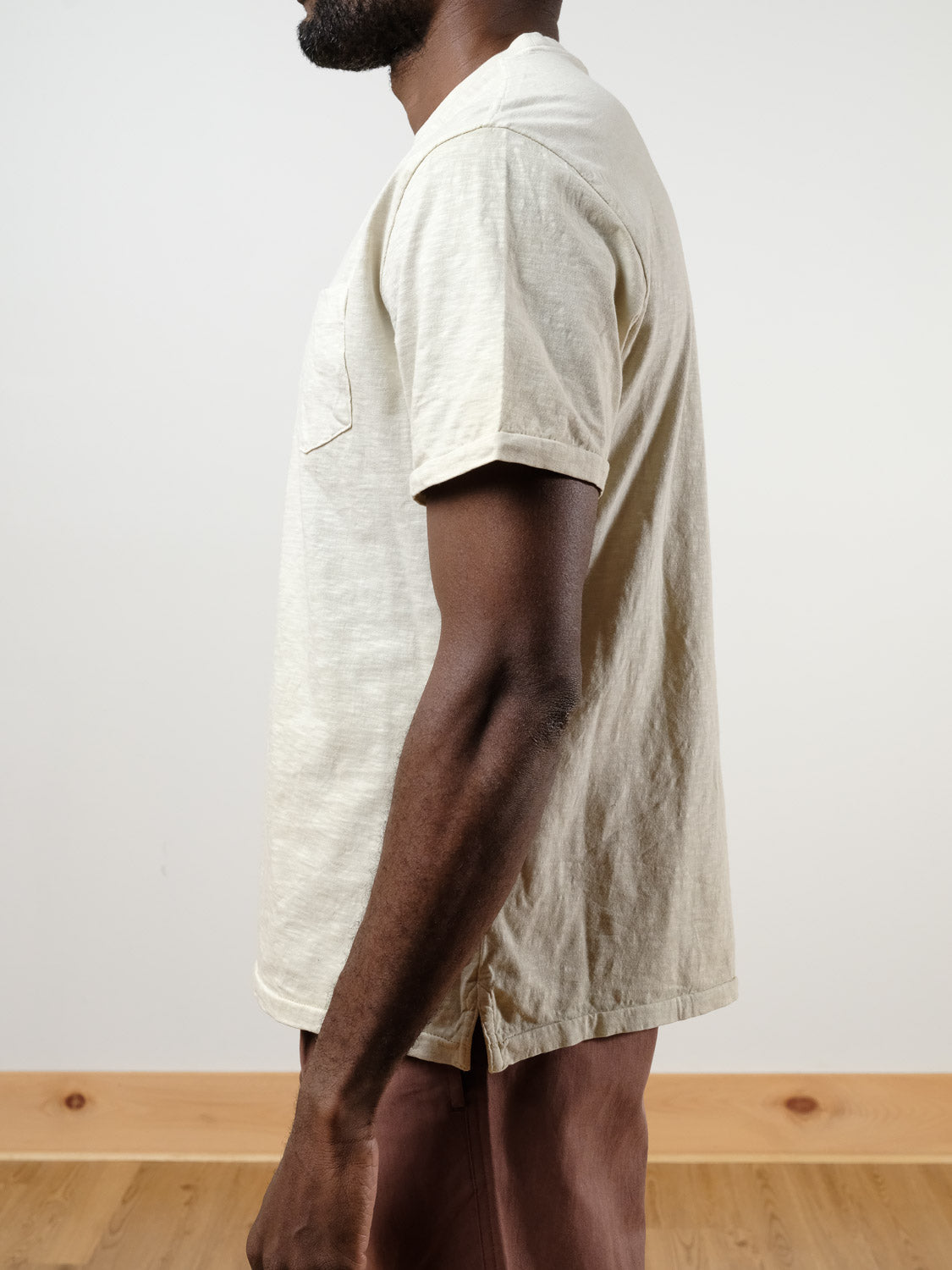A man with a short beard stands sideways, wearing the Rebel Tee by ID Menswear in light beige slub cotton and brown pants. The background features light wood flooring and a plain white wall.