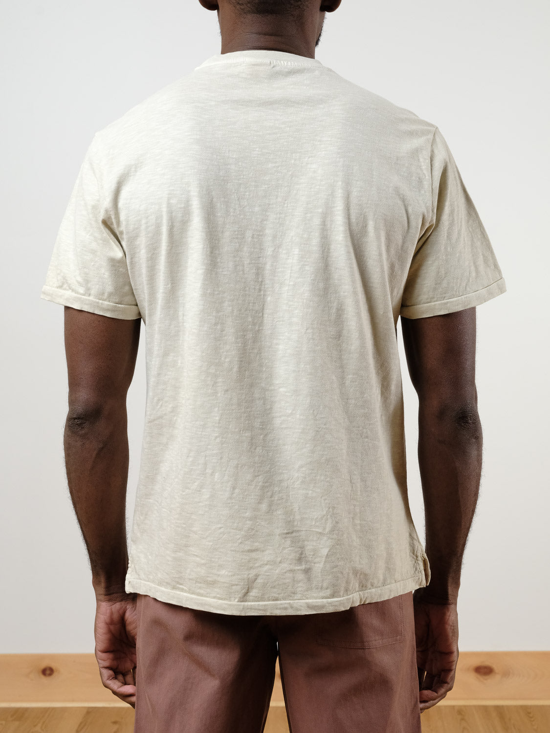 A person stands with their back to the camera, wearing ID Menswear's Rebel Tee in light beige slub cotton and brown pants. The plain, light background and a wooden floor are visible.