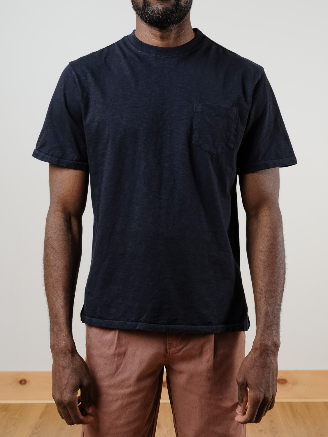 A man wears the ID Menswear Rebel Tee—a relaxed fit, black garment-dyed shirt with a chest pocket—paired with brown pants, standing against a plain light background. His face is not visible.