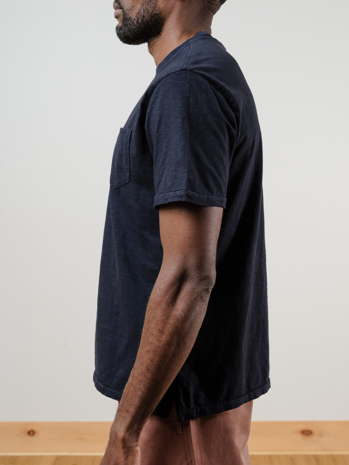 A man with a short beard stands in profile against a plain background, wearing the Rebel Tee by ID Menswear in navy blue and light brown pants.