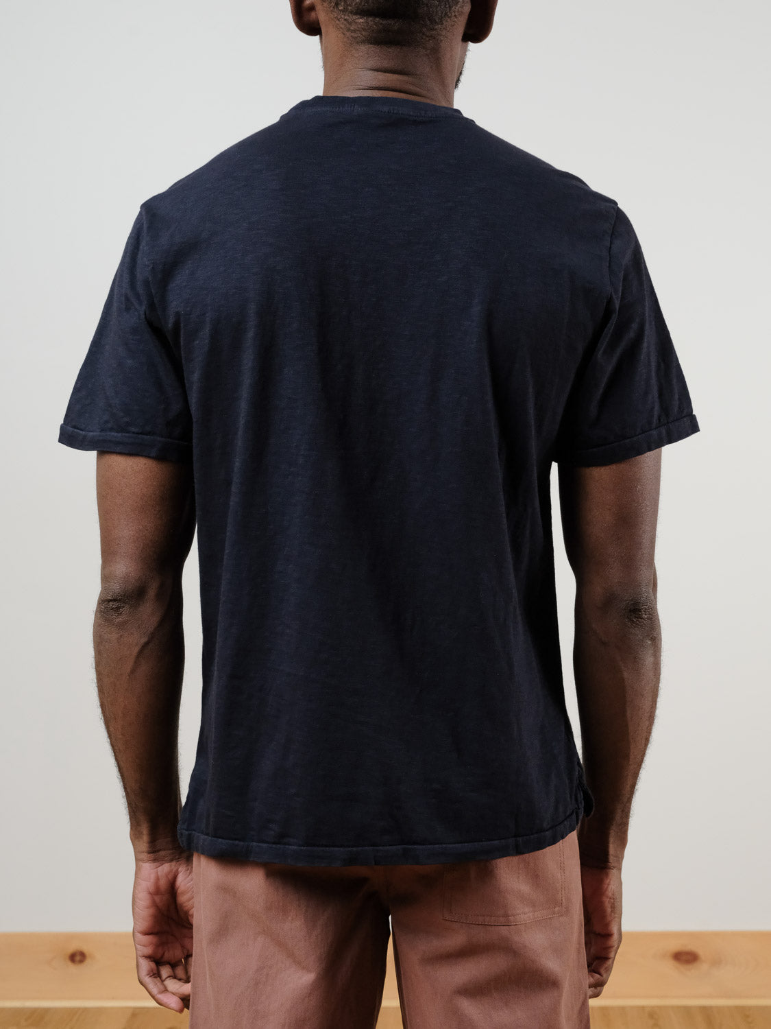 A person faces away from the camera, wearing the Rebel Tee by ID Menswear in dark navy slub cotton with light brown pants against a neutral background.