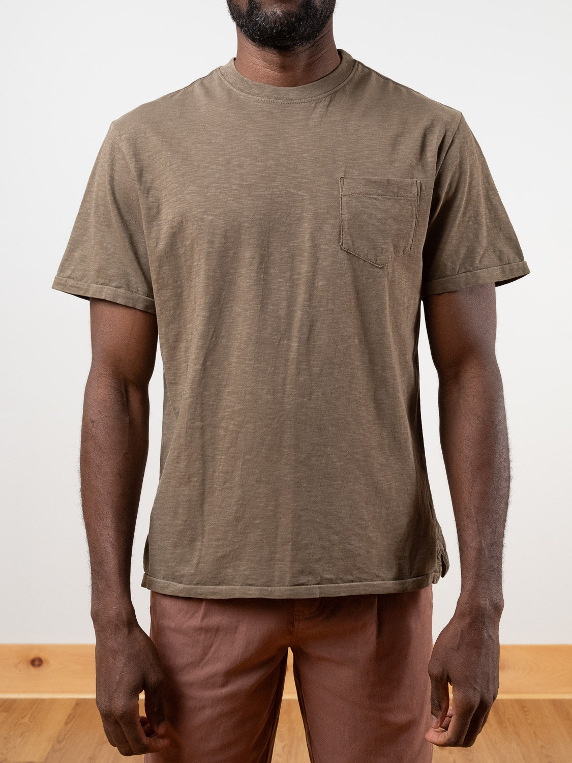 A man wears the Rebel Tee by ID Menswear, a plain brown garment-dyed shirt with a chest pocket, paired with light brown pants, standing against a plain white background.