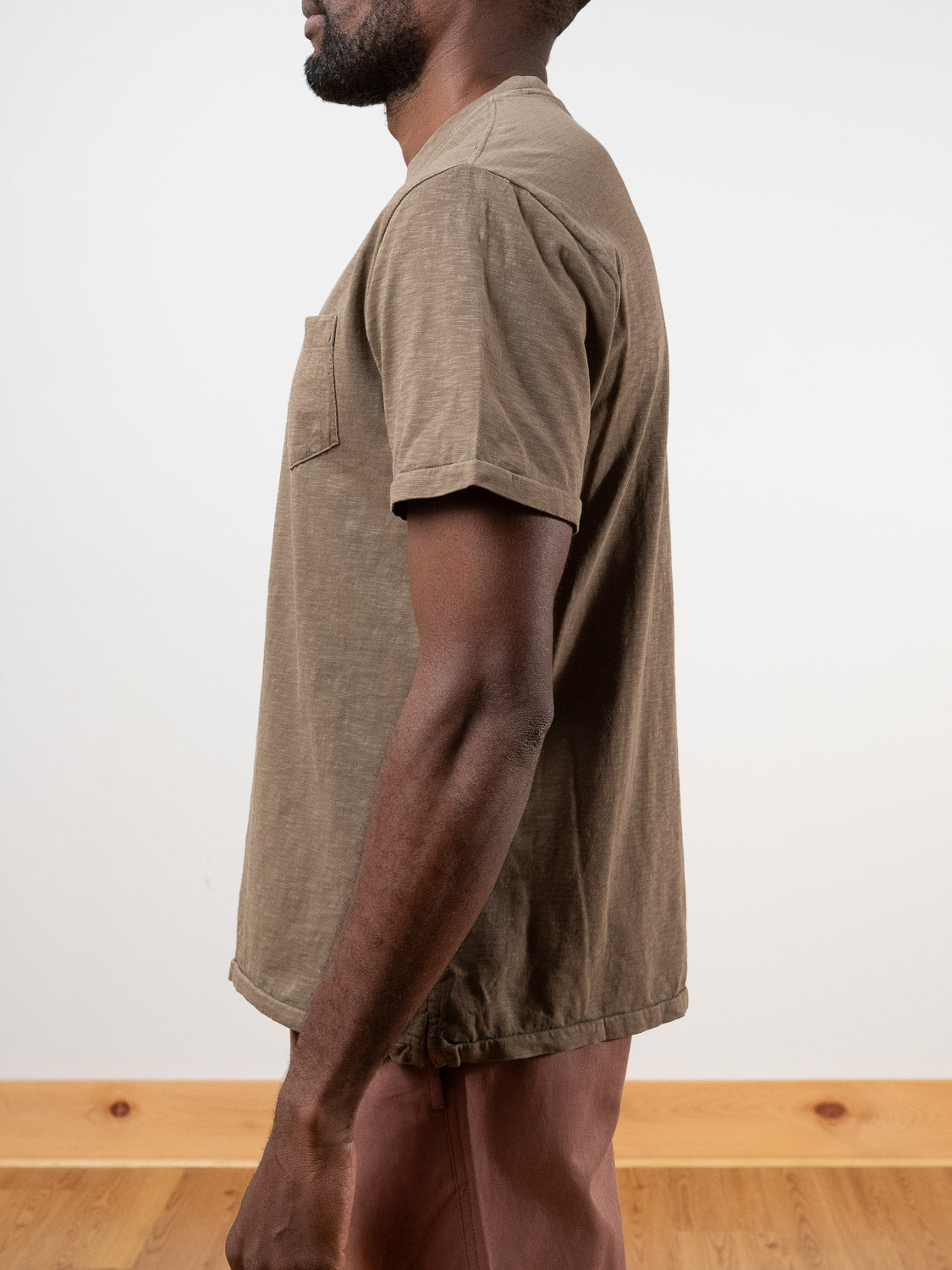 A man stands in profile against a plain background, wearing the Rebel Tee by ID Menswear—a relaxed fit, short-sleeve brown slub cotton t-shirt with a chest pocket. Only the upper half of his body and his light brown pants are visible.