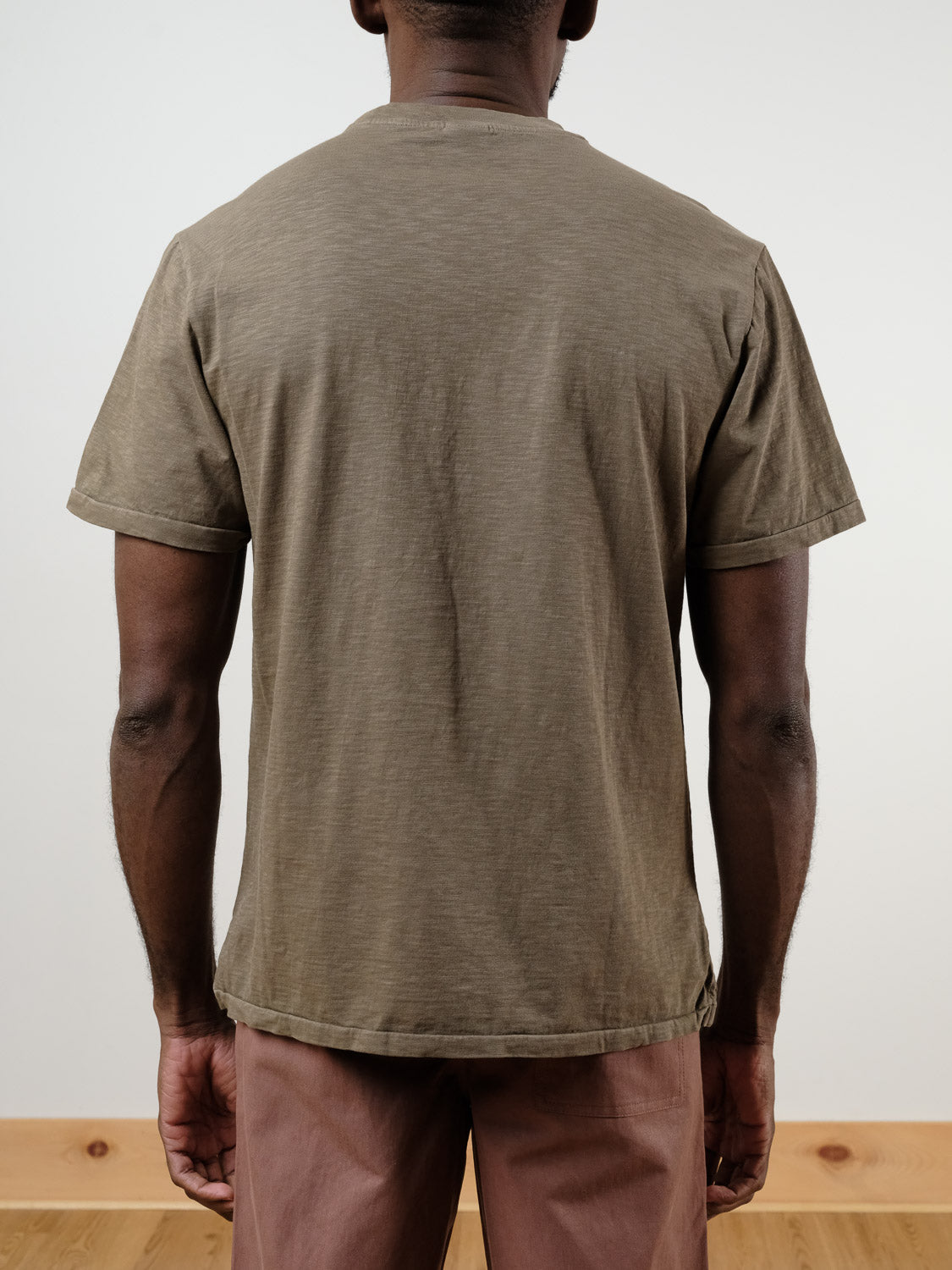 A person faces away from the camera, wearing the short-sleeved Rebel Tee in olive green by ID Menswear with light brown pants, against a plain background and wooden floor.
