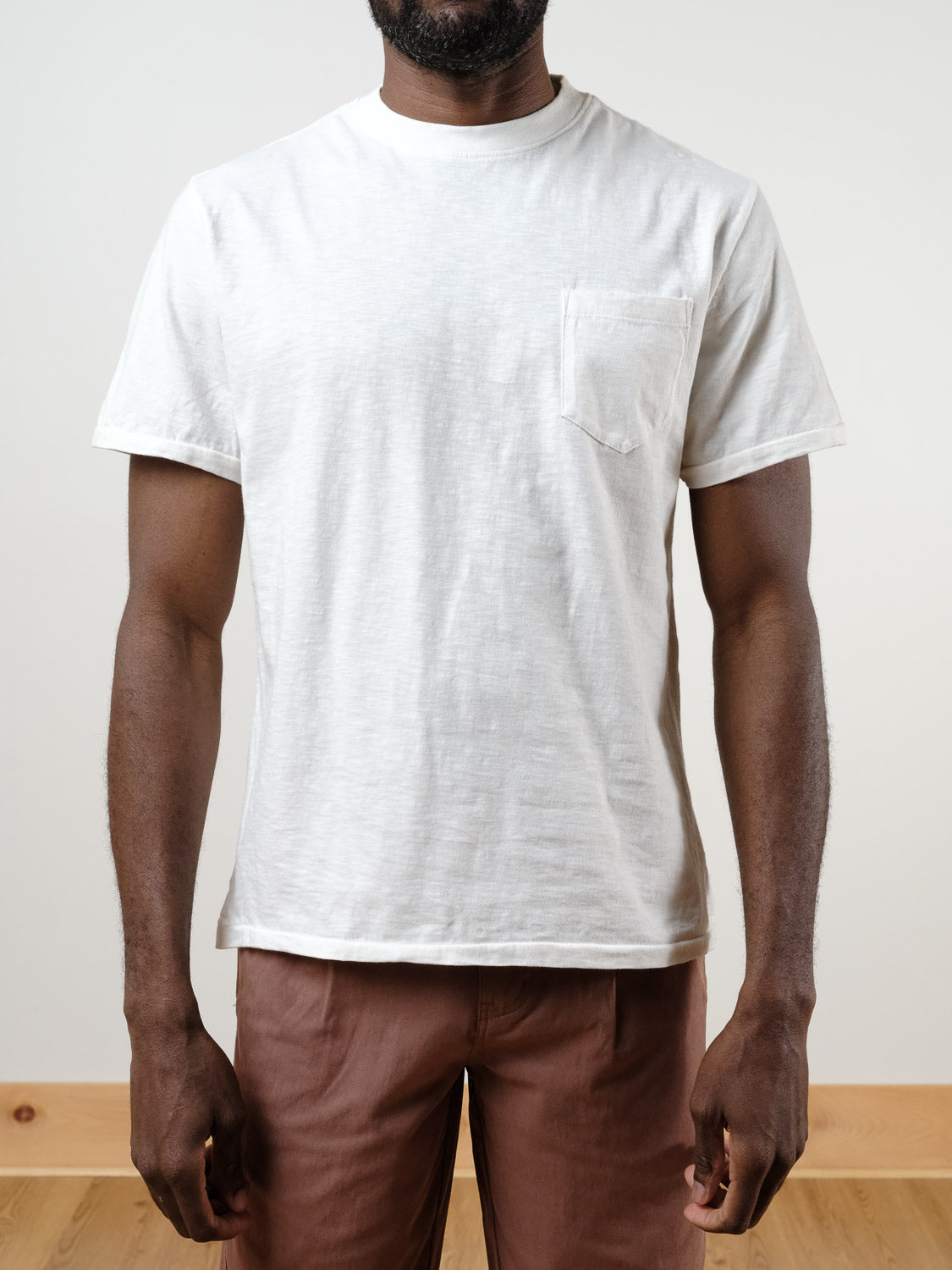 A person wearing the ID Menswear Rebel Tee, a relaxed fit slub cotton t-shirt with a chest pocket, and light brown pants, stands indoors against a neutral background.