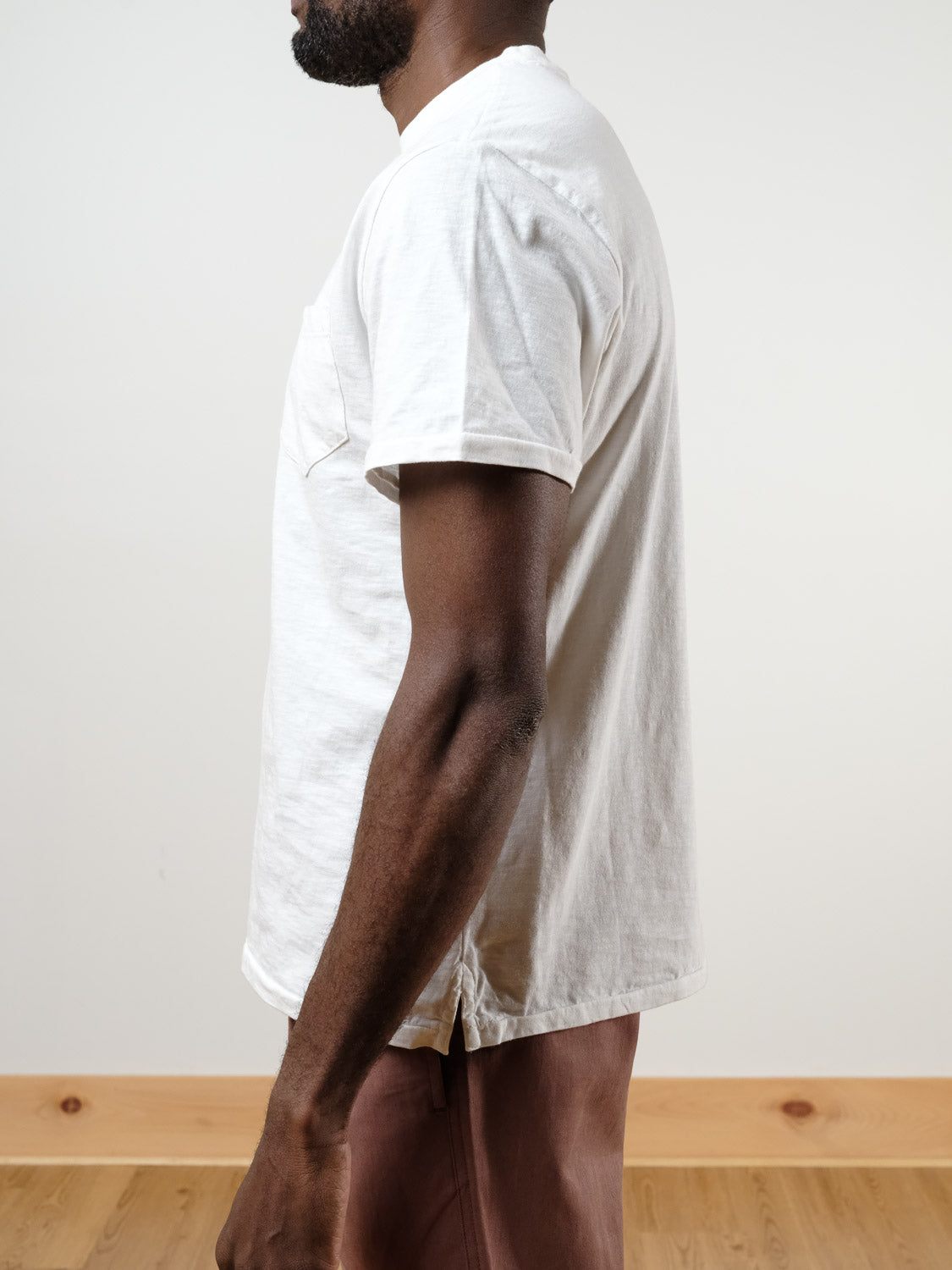 A man stands sideways, showing the left side of his body. He is wearing the Rebel Tee by ID Menswear in white slub cotton and light brown pants. The background is plain with light-colored walls and a wooden floor.