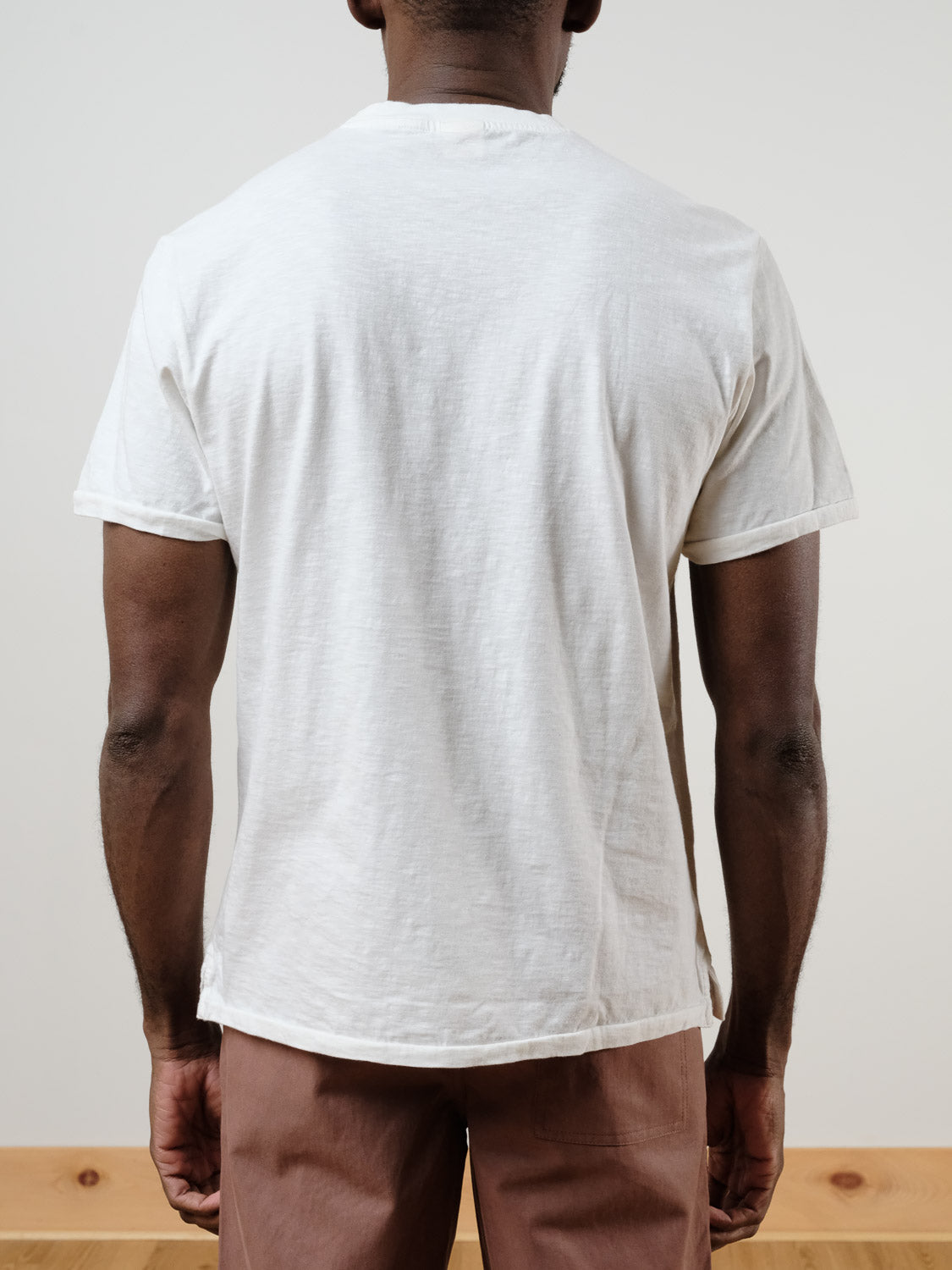 A man faces away from the camera, wearing the ID Menswear Rebel Tee in plain white and light brown pants. The neutral background emphasizes the simple, relaxed look.
