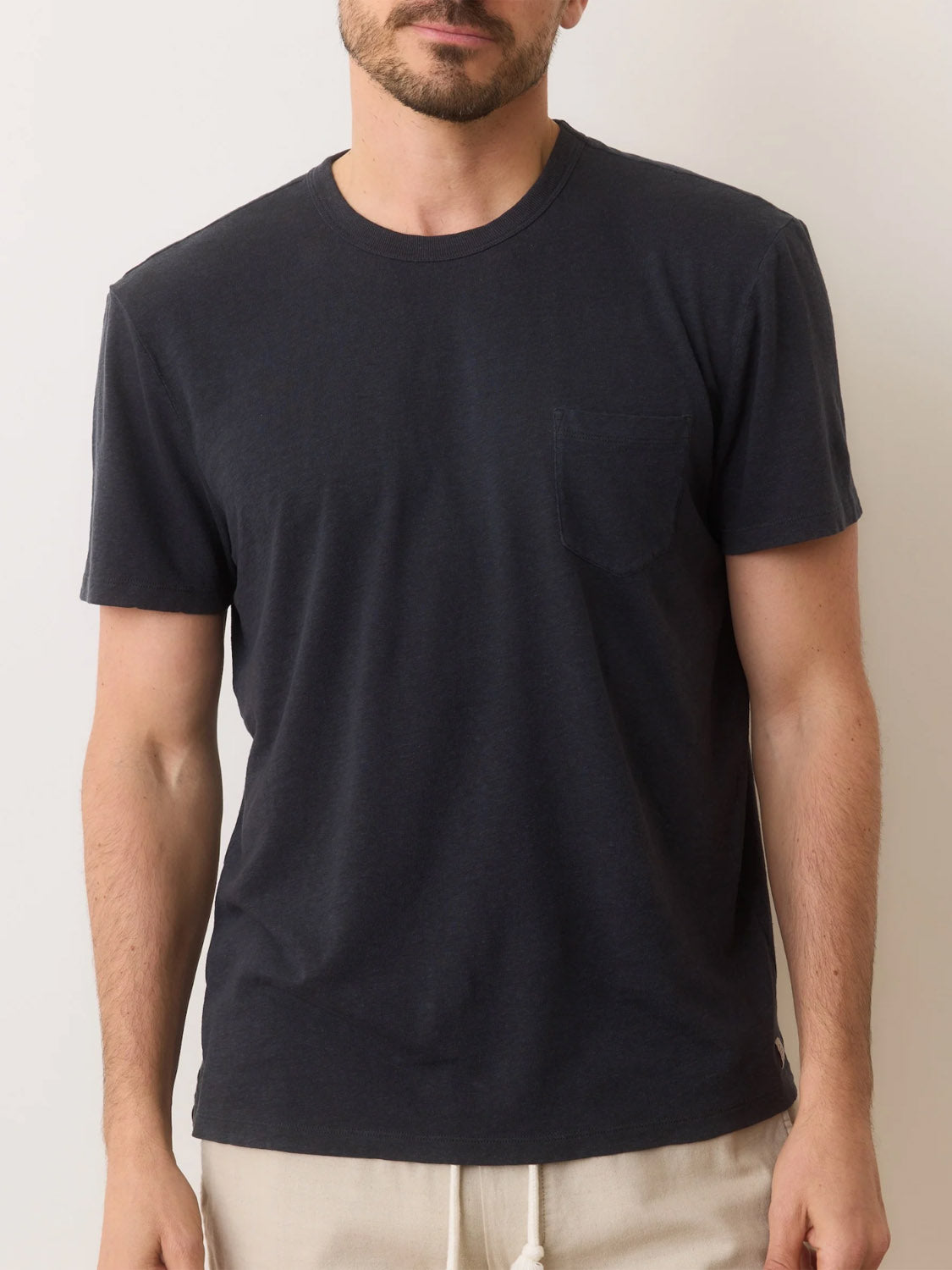 A man wears a Marine Layer Relaxed Hemp Cotton Tee in dark gray, made from breathable fabric with a chest pocket. He pairs it with light-colored pants. Positioned against an off-white wall, the image emphasizes his torso and arms, with his head partially visible.