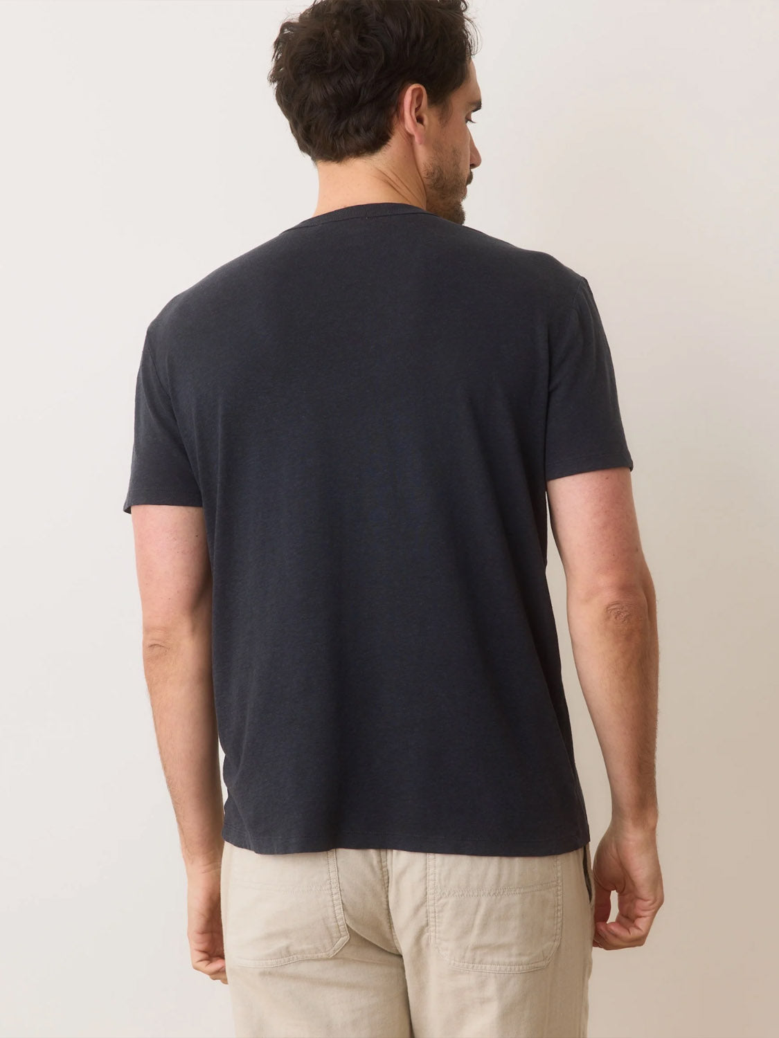 A man is seen from the back wearing a Marine Layer Relaxed Hemp Cotton Tee in dark gray and beige pants, against a light background. His head slightly turned left emphasizes the breathable fabric known for ultimate comfort provided by Marine Layer.