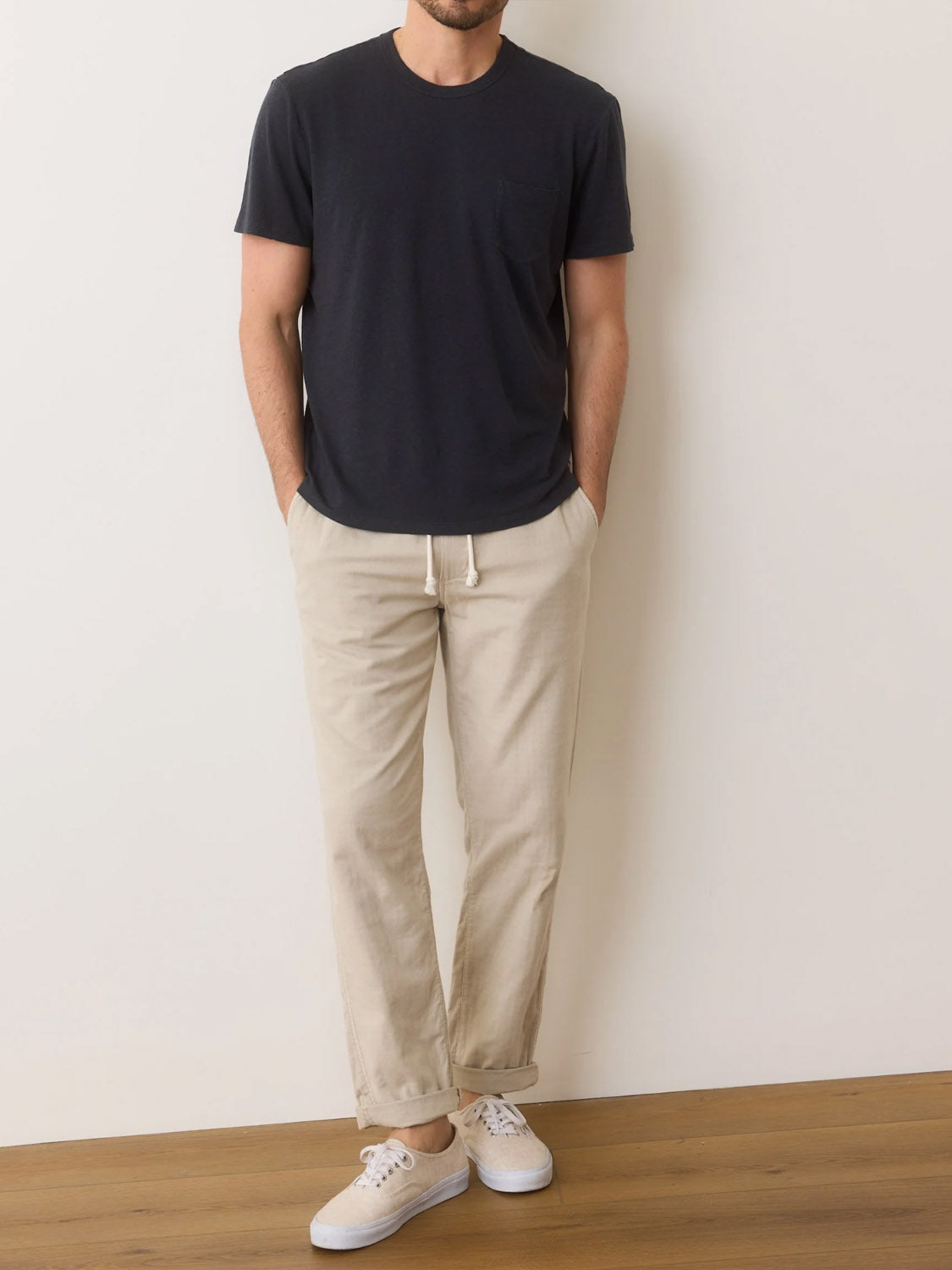 A person stands against a plain wall, wearing a comfortable Marine Layer Relaxed Hemp Cotton Tee in black, beige drawstring pants, and white sneakers. Their hands rest in their pockets, with wooden flooring visible beneath them.