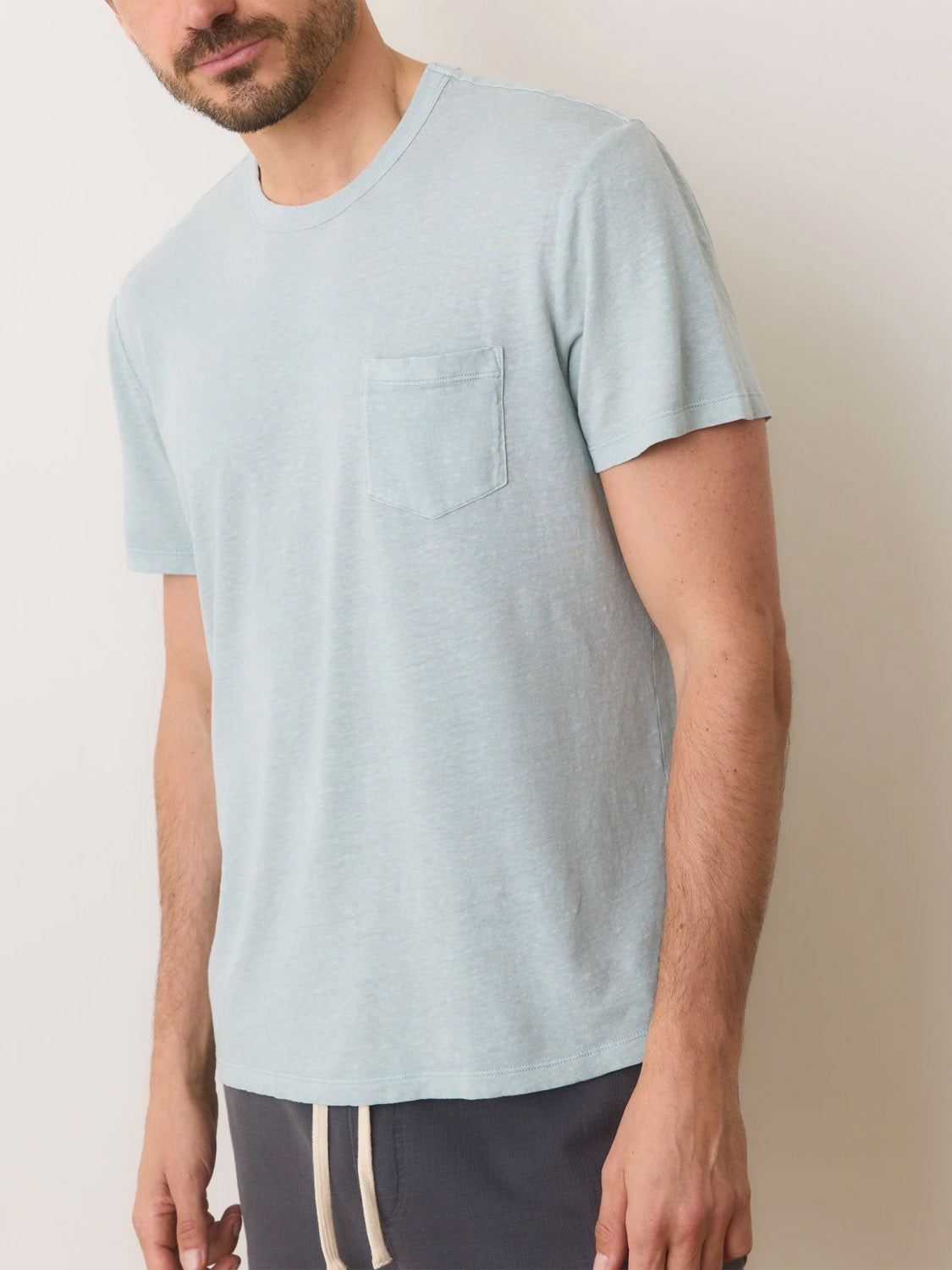 A man in a Marine Layer Relaxed Hemp Cotton Tee in light blue, featuring a pocket, pairs it with dark shorts. Standing against a plain white backdrop, he displays casual ease, with his face partly visible and arms relaxed at his sides.