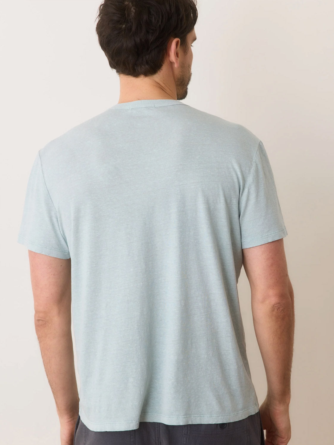 A person with short dark hair stands with their back to the camera, wearing a Marine Layer Relaxed Hemp Cotton Tee in light gray and dark pants. They are set against a plain, neutral wall.