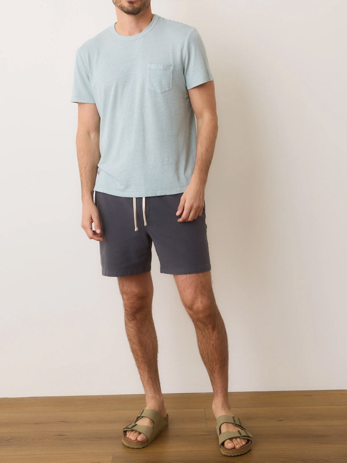 Man in a Marine Layer Relaxed Hemp Cotton Tee of light blue, teamed with dark shorts and beige sandals, stands on a wooden floor against a white wall. Face is not visible.