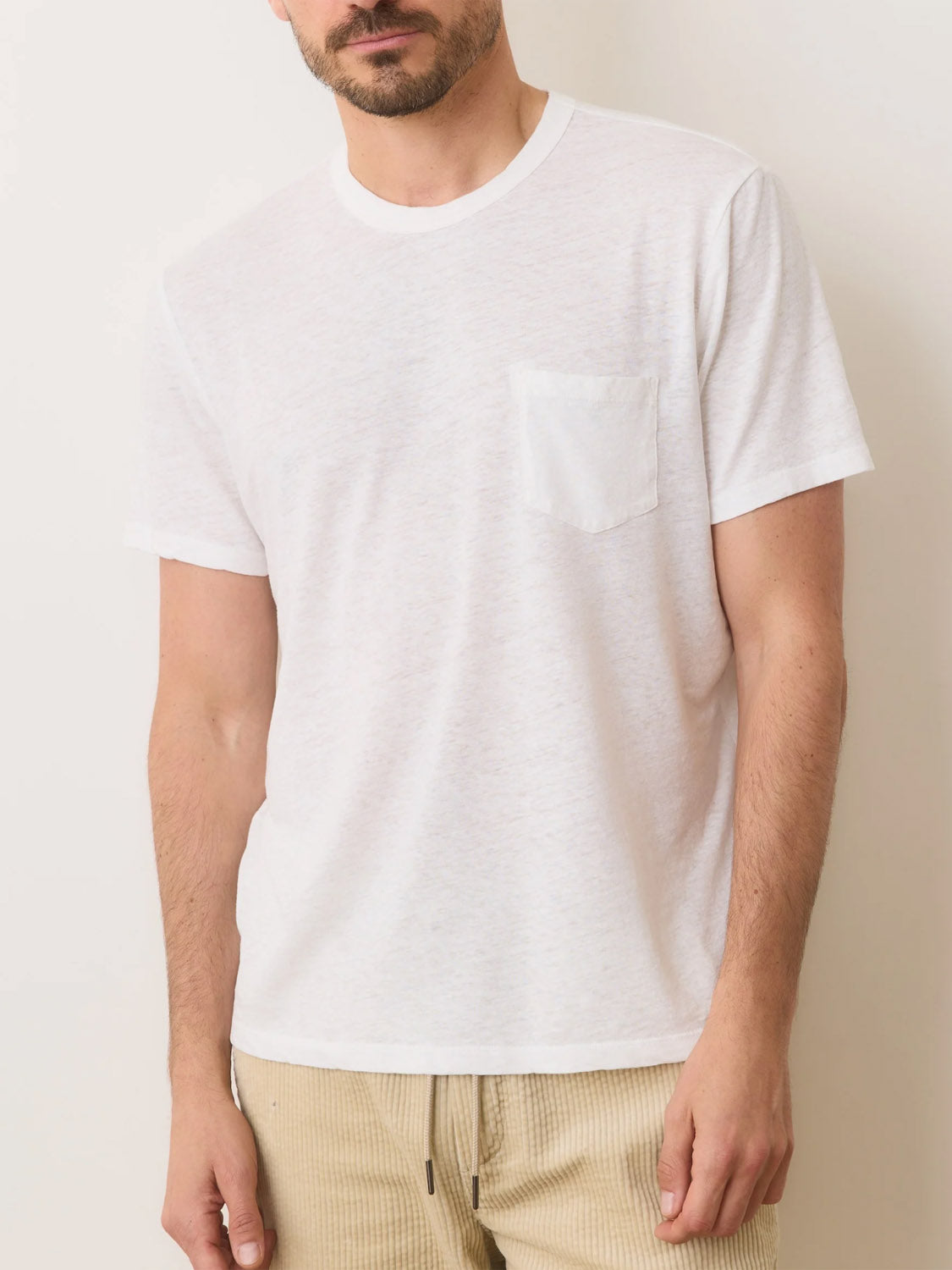 A person is wearing a Marine Layer Relaxed Hemp Cotton Tee in plain white, featuring a left-side pocket, paired with beige pants. The figures head is partially cropped out against a simple background.