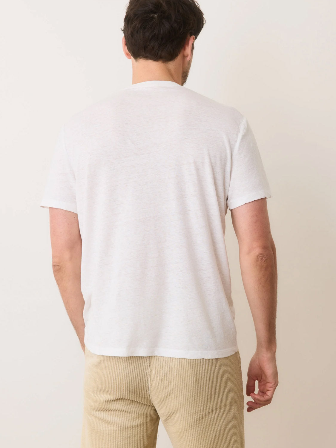 A man with short dark hair is facing away, wearing a Marine Layer Relaxed Hemp Cotton Tee and beige corduroy pants against a simple, light-colored wall.