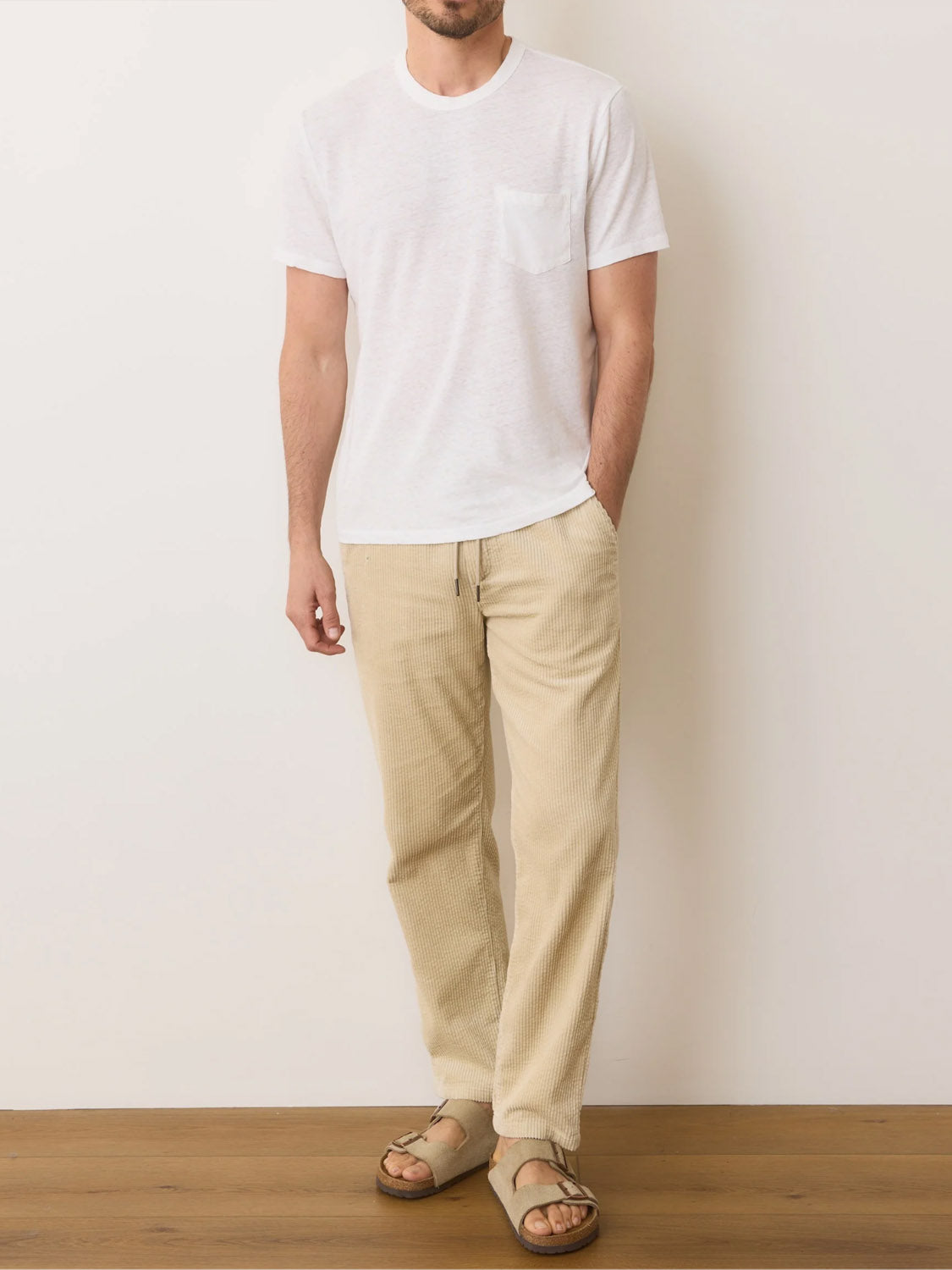 A man in a Marine Layer Relaxed Hemp Cotton Tee stands against a plain wall and wooden floor, dressed in beige textured pants and brown sandals. His hands rest in his pockets, obscuring his face.