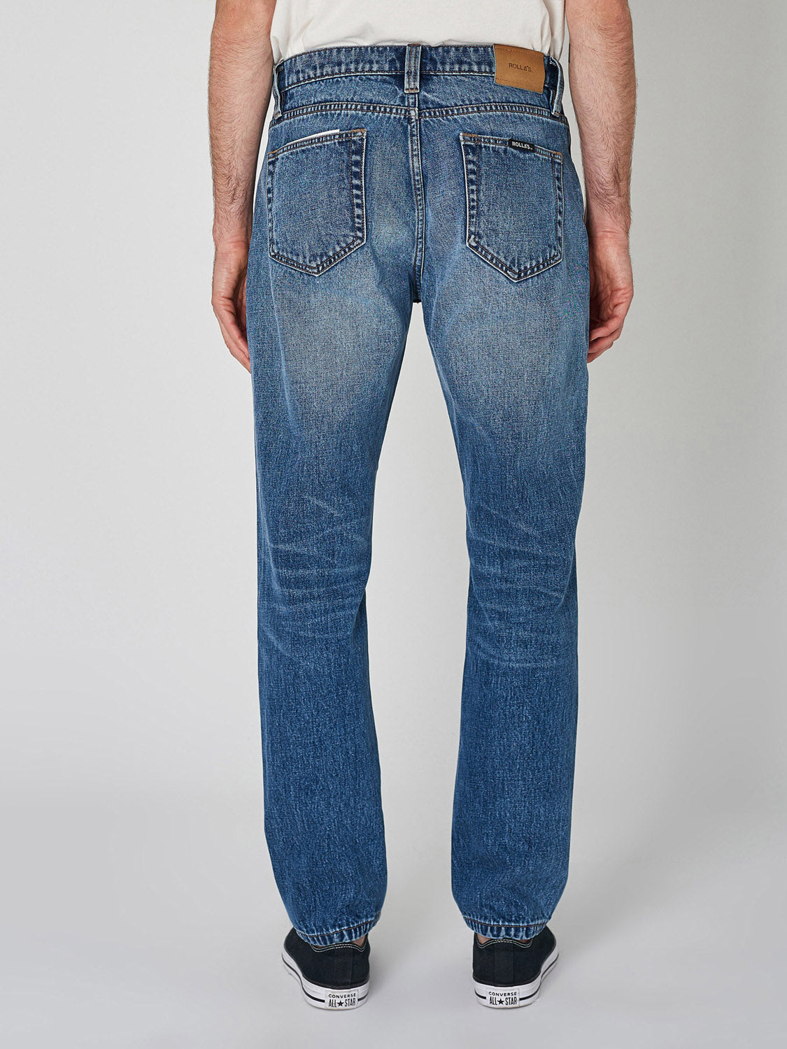A person in a white shirt, Rollas Relaxo Hemp Blues Denim, and black sneakers stands facing away from the camera, showcasing the back view of the jeans and shoes against a plain background.