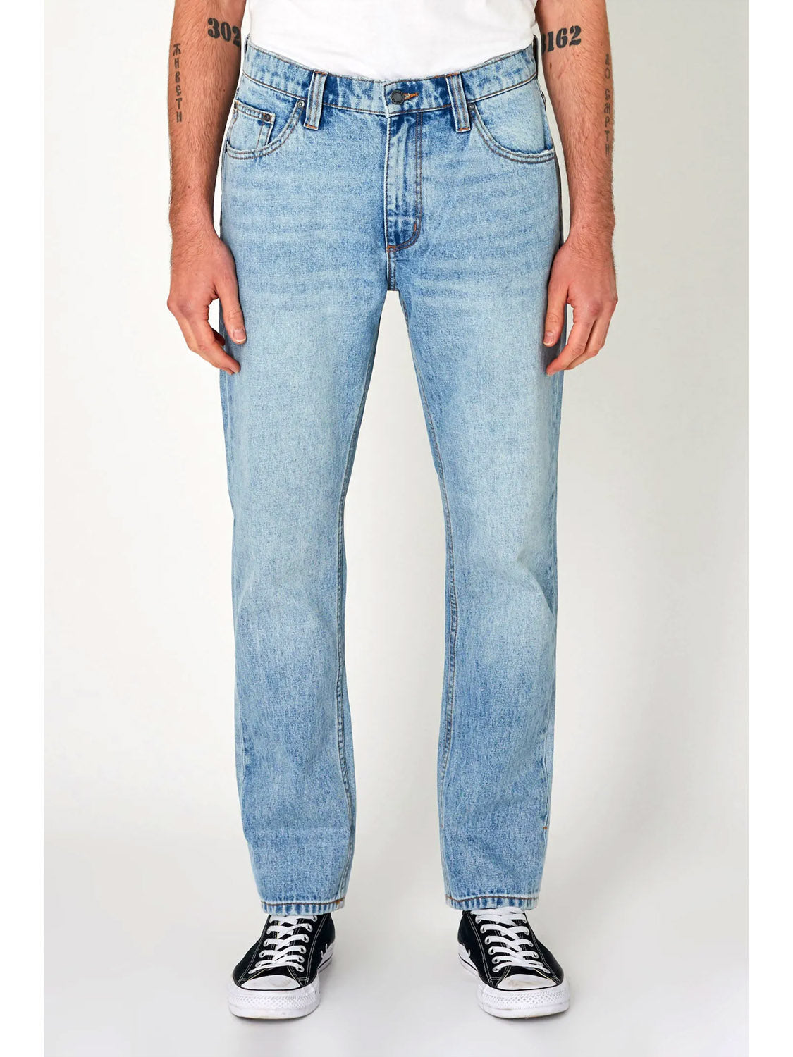 A person wears Rollas Relaxo Original Stone Denim in light blue with a white T-shirt and black-and-white sneakers, standing against a plain white background. Tattoos are visible on their arms.