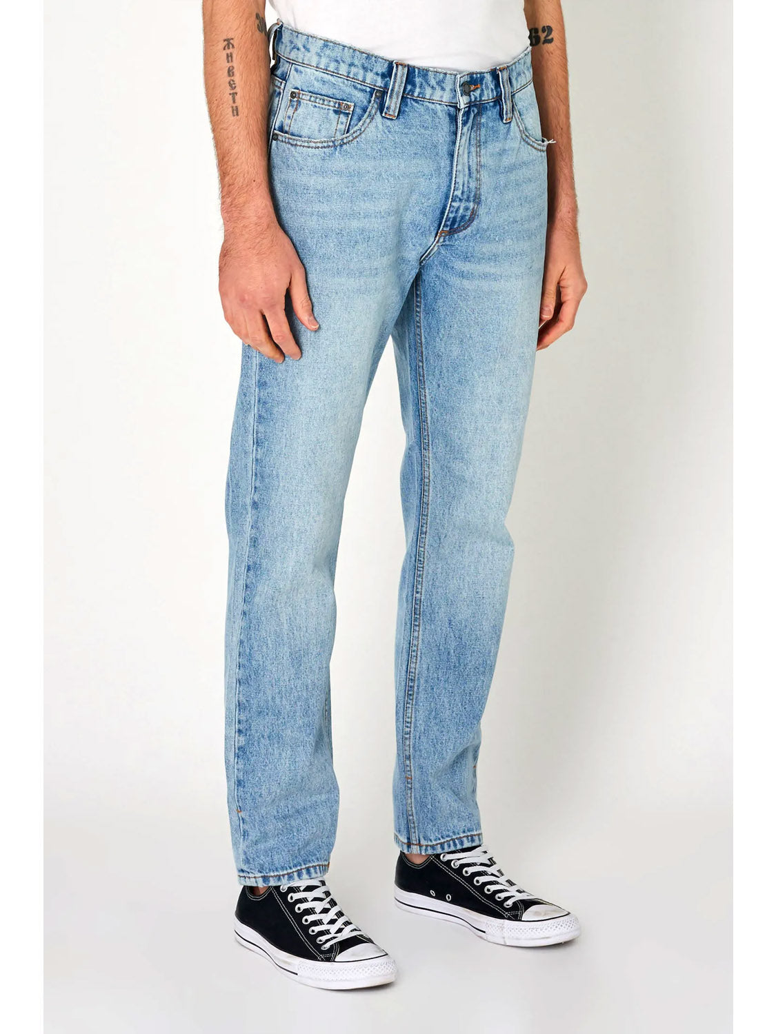 A person wearing Rollas Relaxo Original Stone Denim jeans, a white t-shirt, and black low-top Converse sneakers stands against a plain white background. Tattoos are visible on the person’s arms.