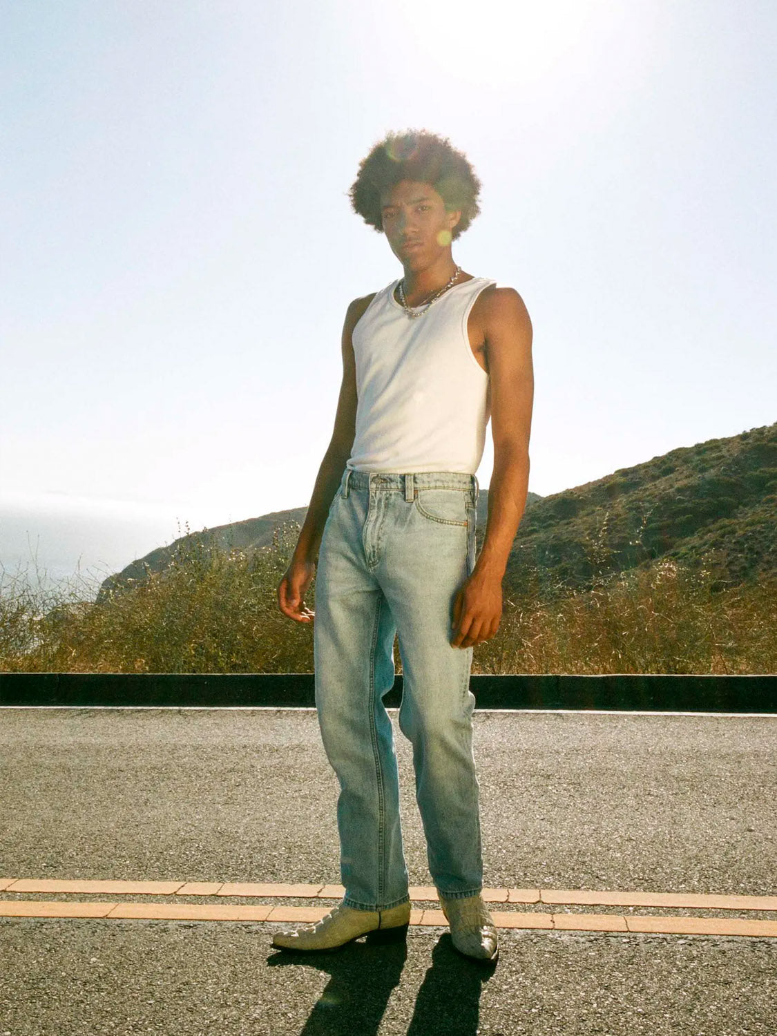 Wearing a white tank top, white shoes, and Rollas Relaxo Original Stone Denim jeans, a person stands on a sunlit road with hills and dry grass behind them. Sunlight creates a soft glow around their head.