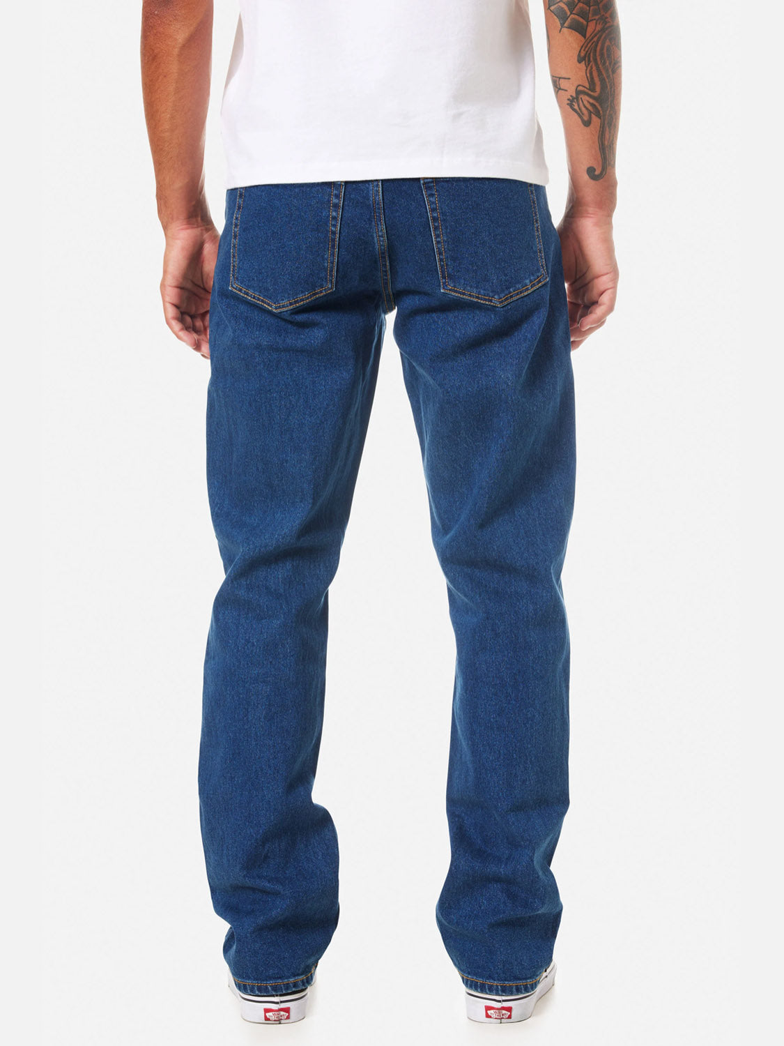 A man stands facing away, wearing a white T-shirt, white sneakers, and Katin Ren 57 Classic Straight Pants with 5-pocket styling. A tattoo is visible on his right arm.