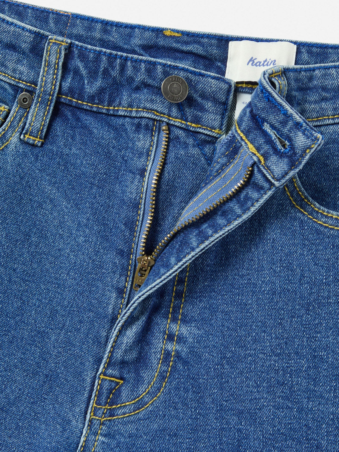 Close-up of Katin Ren 57 Classic Straight Pants in blue denim, featuring a partially unzipped brass zipper, metal waistband button, yellow stitching, 5-pocket styling, and a visible fabric tag with the Katin brand name.