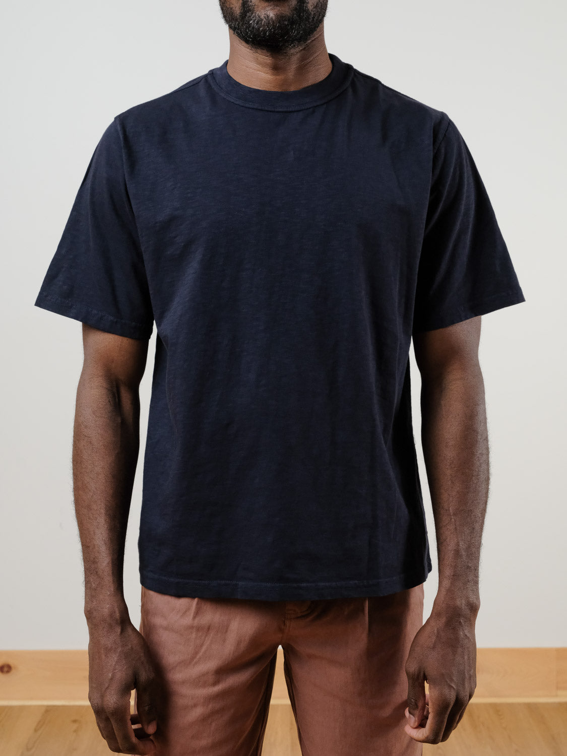 A man stands against a plain light background wearing the Renegade Tee by ID Menswear in navy blue, paired with light brown pants. His arms are relaxed at his sides and his head is cropped out of the frame.