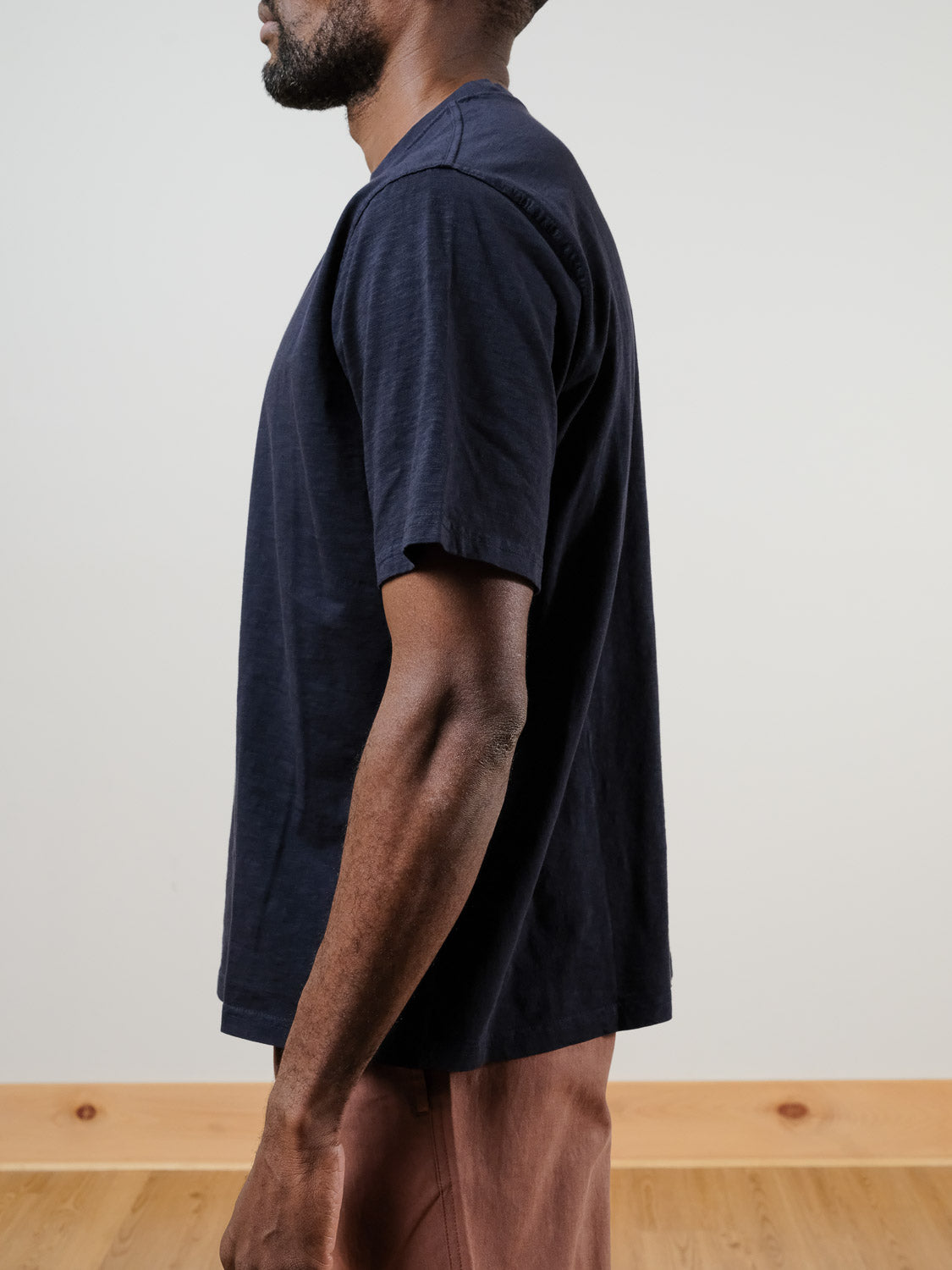 A man stands in profile against a plain background, wearing the dark navy Renegade Tee by ID Menswear (100% cotton) paired with light brown pants. The image highlights his upper body and arm.