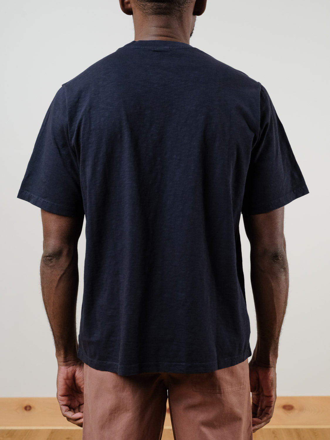 A person stands with their back to the camera, wearing the ID Menswear Renegade Tee in plain navy blue 100% cotton and light brown pants against a neutral background.