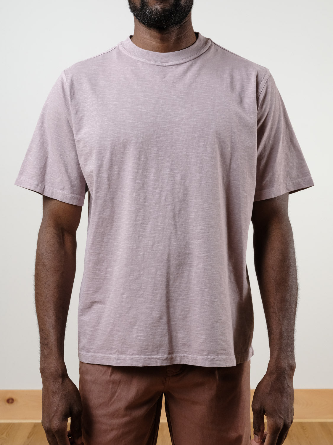 A person stands facing forward in a light mauve Renegade Tee by ID Menswear and brown pants. The 100% cotton, short-sleeve t-shirt has a boxy fit, and the plain background keeps the person’s face out of view.