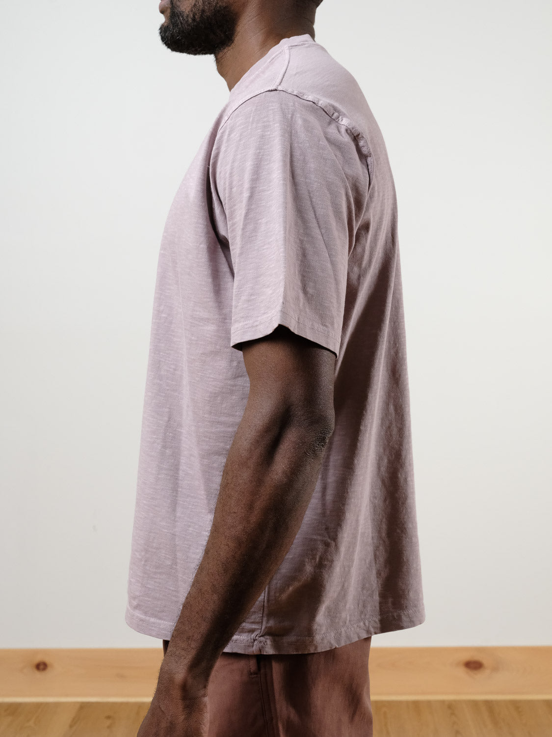 A man stands in profile against a plain background, wearing the Renegade Tee by ID Menswear in light mauve with matching pants, highlighting the loose fit and side view of his torso and arm.