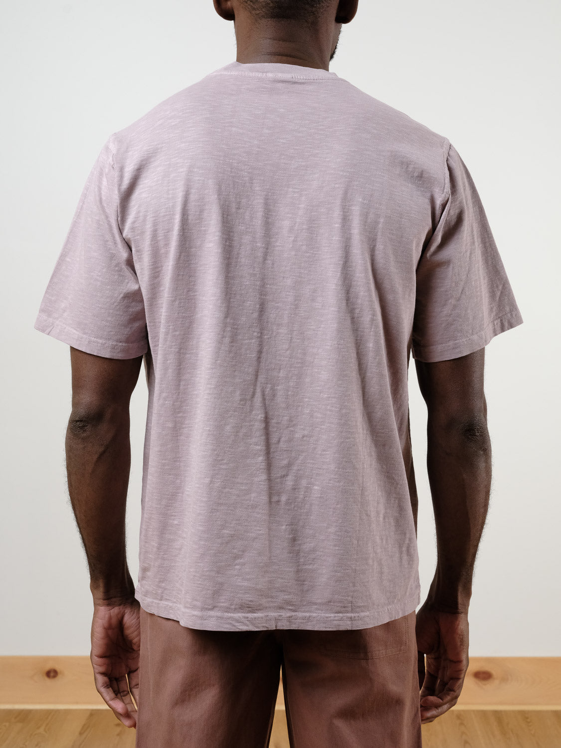 A person faces away from the camera, wearing the light mauve Renegade Tee by ID Menswear and matching pants, with hands relaxed by their sides against a plain background.