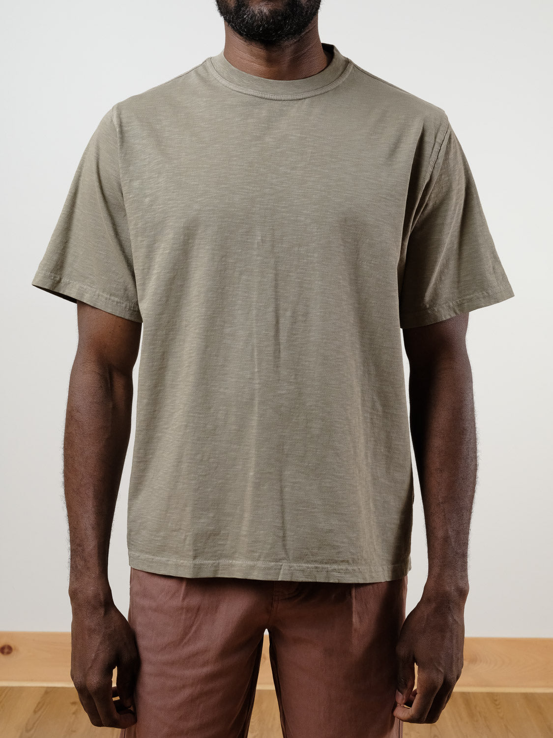 A man models the ID Menswear Renegade Tee, a loose-fit 100% cotton t-shirt in light olive green, paired with brown pants. He stands against a plain, light background with his face not shown.