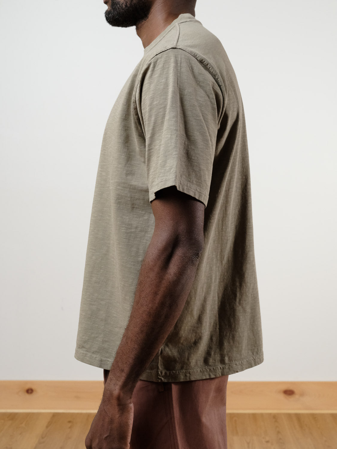 A person stands in profile wearing the ID Menswear Renegade Tee, a loose-fitting, short-sleeve, garment-dyed t-shirt in olive green, paired with brown pants. The plain white wall and wooden floor serve as the background.