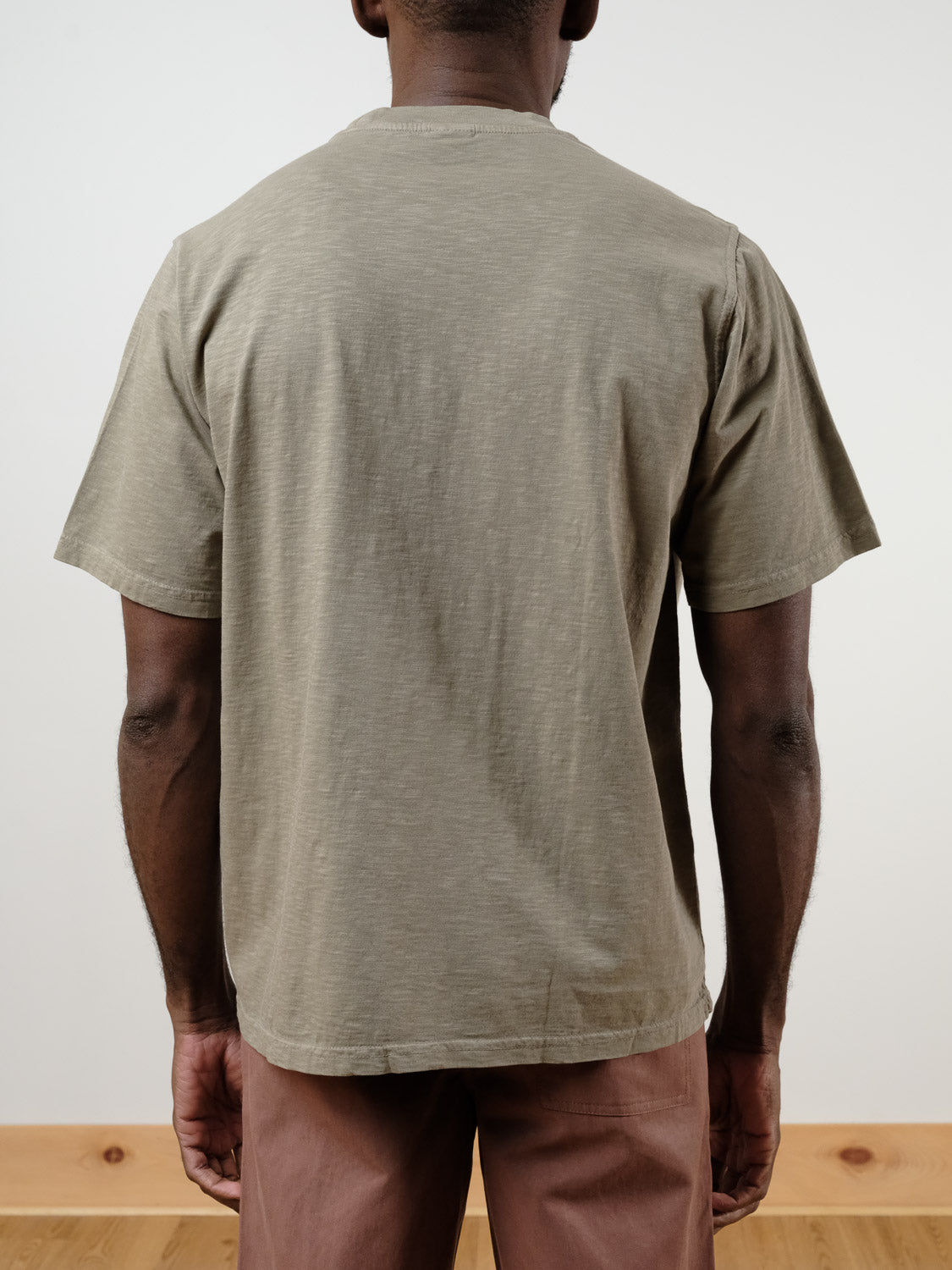 A person faces away from the camera, wearing the Renegade Tee by ID Menswear—a loose, garment-dyed, boxy t-shirt in light brown—paired with brown pants against a plain light background.