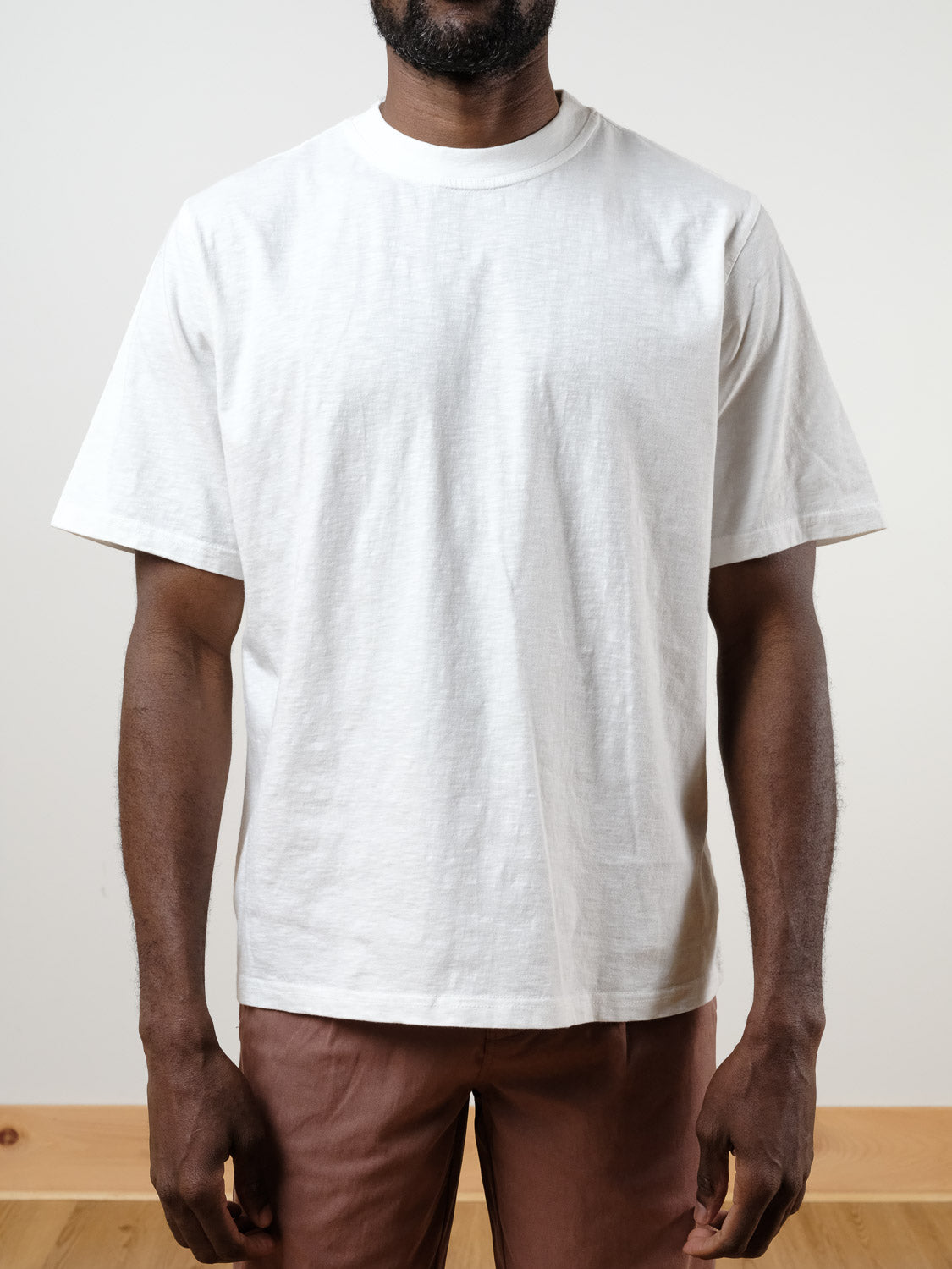 A man wearing the ID Menswear Renegade Tee, a plain white 100% cotton t-shirt, and brown pants stands against a light background, shown from shoulders to thighs.