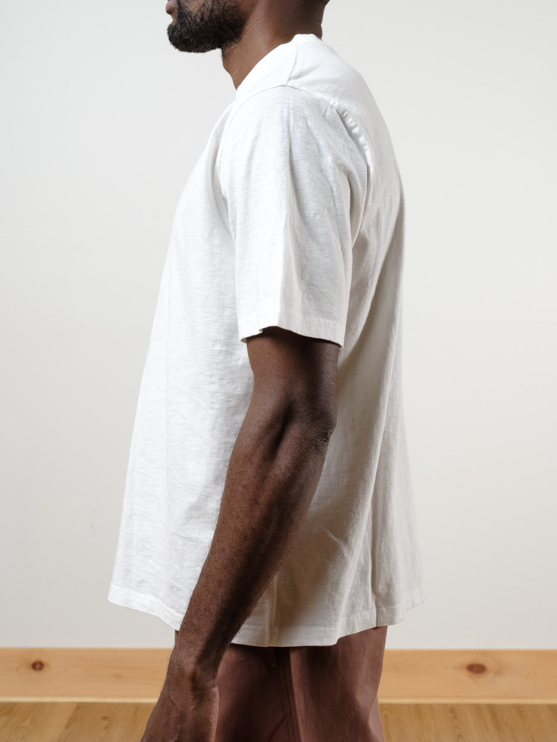 A person stands in profile wearing the boxy Renegade Tee by ID Menswear, made from 100% cotton, paired with brown pants. The background is light with a wooden floor, and the person's face is partially out of view.