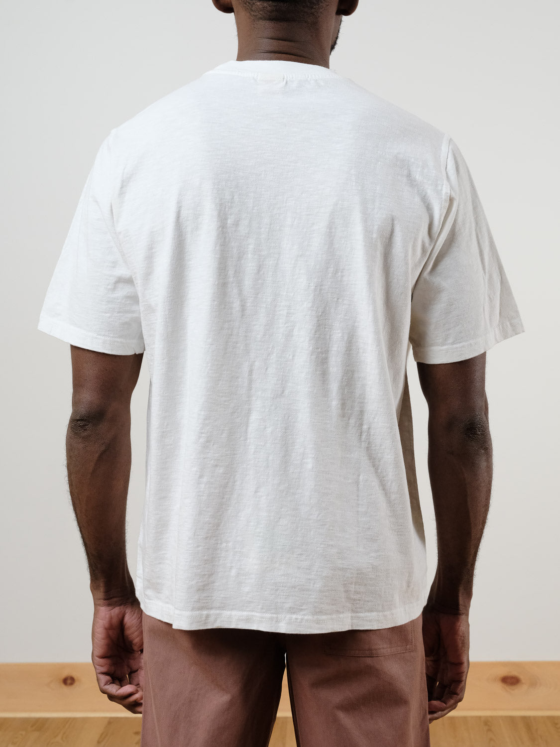 A person stands indoors facing away, wearing the Renegade Tee by ID Menswear—a boxy 100% cotton plain white t-shirt—and light brown pants. The scene has a neutral background and a wooden floor.