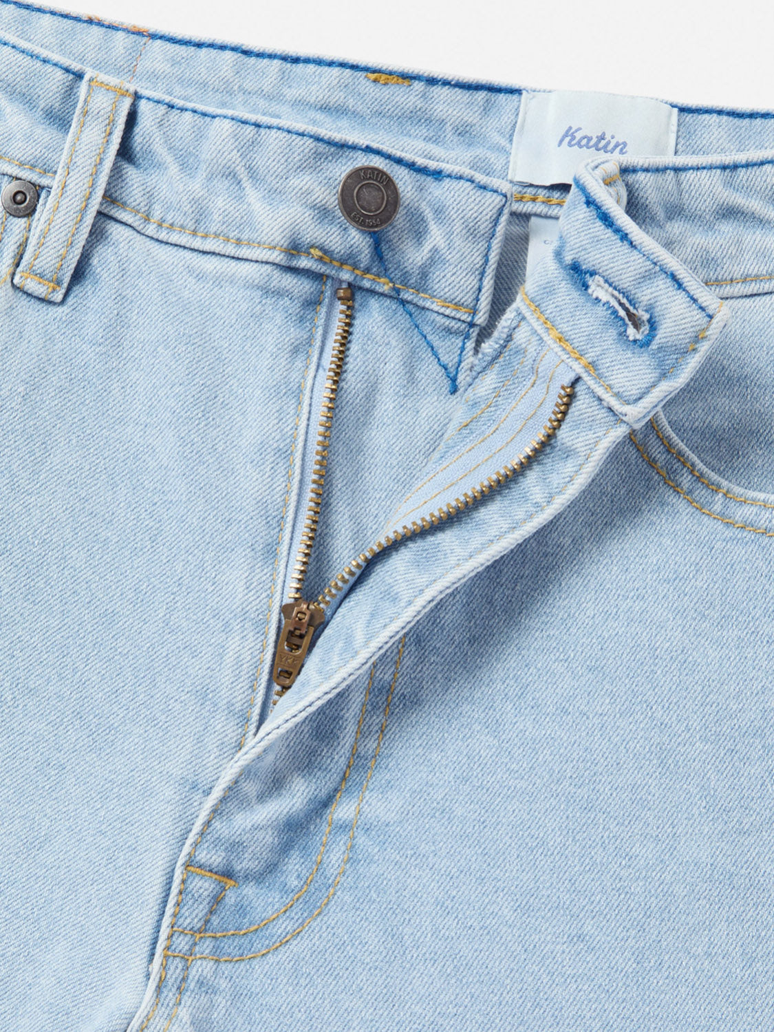 Close-up of Katin Ren 57 Classic Straight Pants in light blue denim, featuring classic 5-pocket styling, a partially open bronze zipper, metal waistband button, and a white label with blue Katin text.