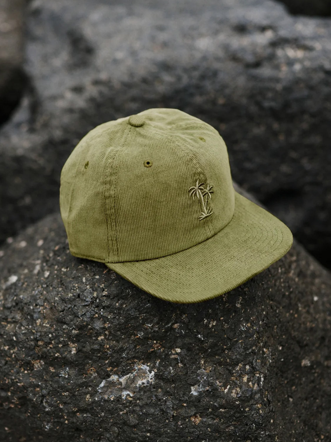 The Katin Retreat Hat, in olive green corduroy with an embroidered palm tree and snapback closure, is shown resting on a rugged dark rock surface.