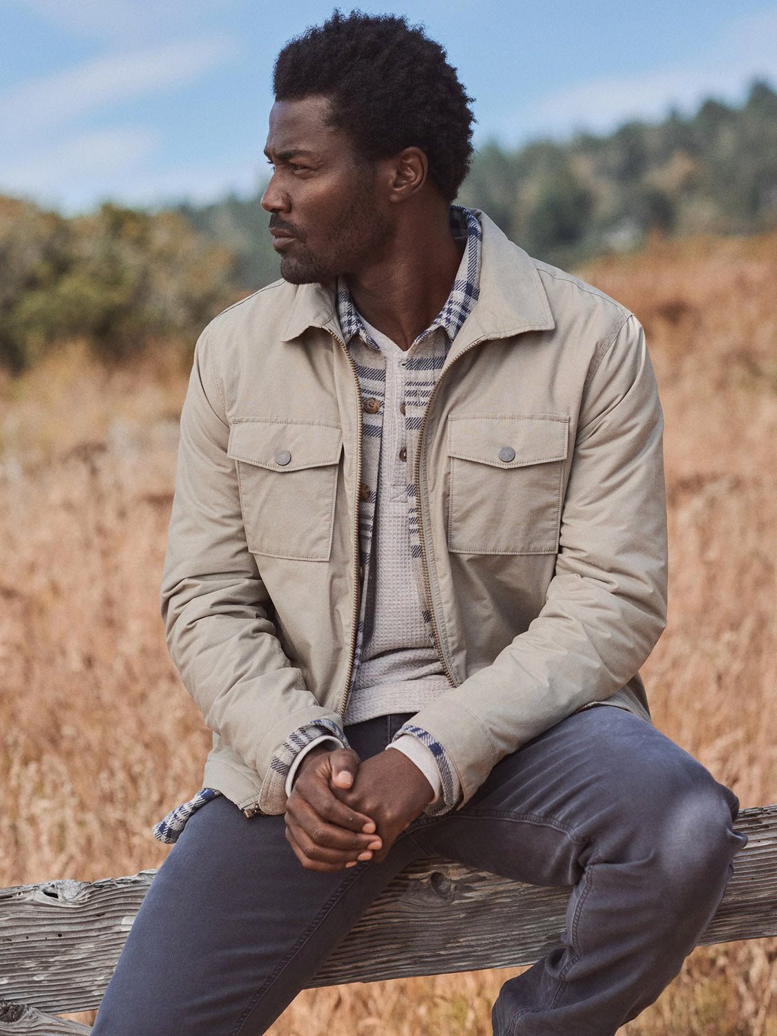 A man sits outdoors on a wooden fence, wearing the Marine Layer Reynolds Dry Wax Trucker Jacket over a plaid shirt and dark pants, looking left with a dry grassy field and trees in the background.