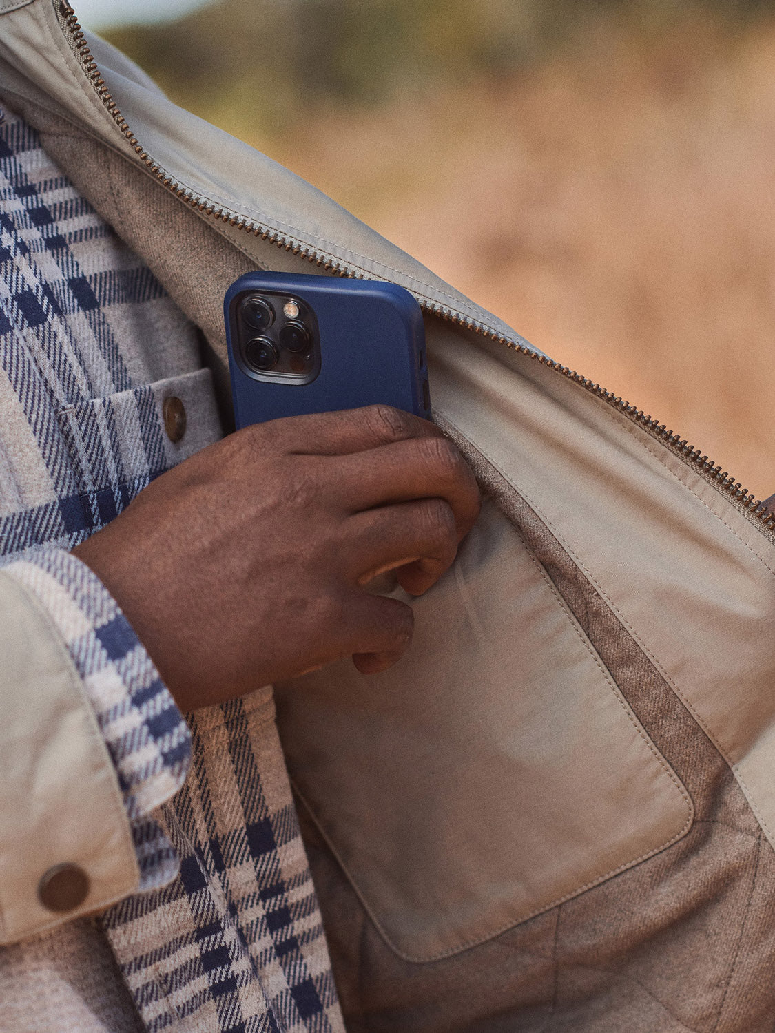 A person wearing a plaid shirt and the Marine Layer Reynolds Dry Wax Trucker Jacket puts a blue smartphone into the jacket's inner pocket. The blurred background hints at an outdoor setting.