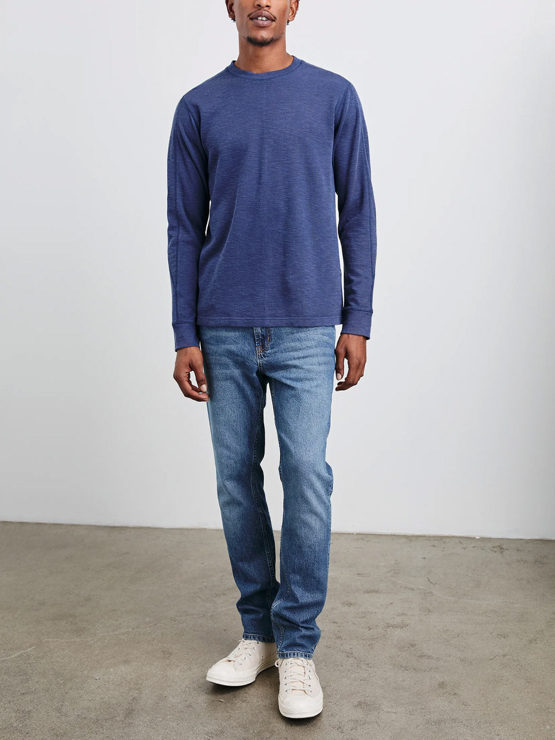 A person in a Rails Rheese Long Sleeve Thermal and crewneck jeans stands on a textured concrete floor. They wear white sneakers, epitomizing elevated essentials against a plain light-colored wall.