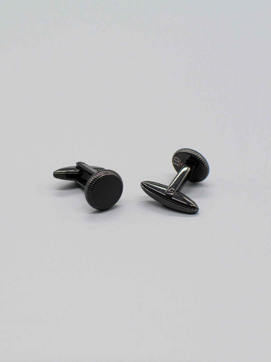 Two Black Gold Plated Steel Enamel Cufflinks by ID Menswear rest on a gray surface. Each has a round face with a polished design and are made from black gold-plated steel. Slightly tilted, they highlight their sleek elegance and bullet back mechanism.