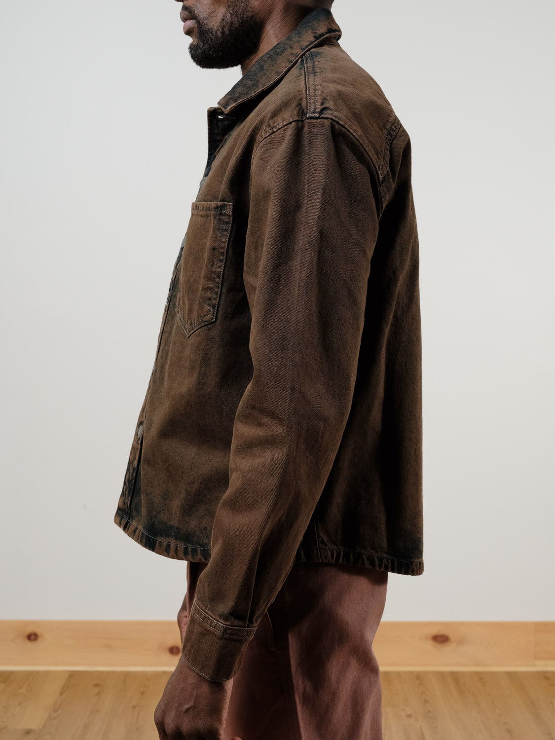A man stands sideways against a plain light-colored wall and wooden floor, wearing the !SOLID Rip Vintage Washed Denim Jacket and brown pants.