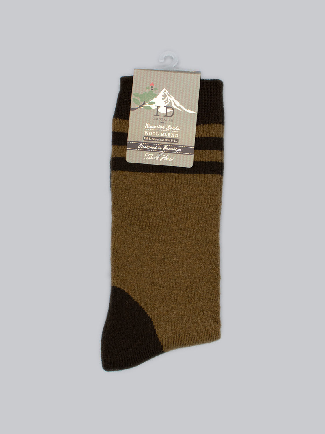 A single Rockies Merino Wool sock from ID Menswear is shown vertically against a light gray background. Predominantly brown with darker stripes and toes for warmth and durability, it features a mountain-logo cardboard label at the top with product details.