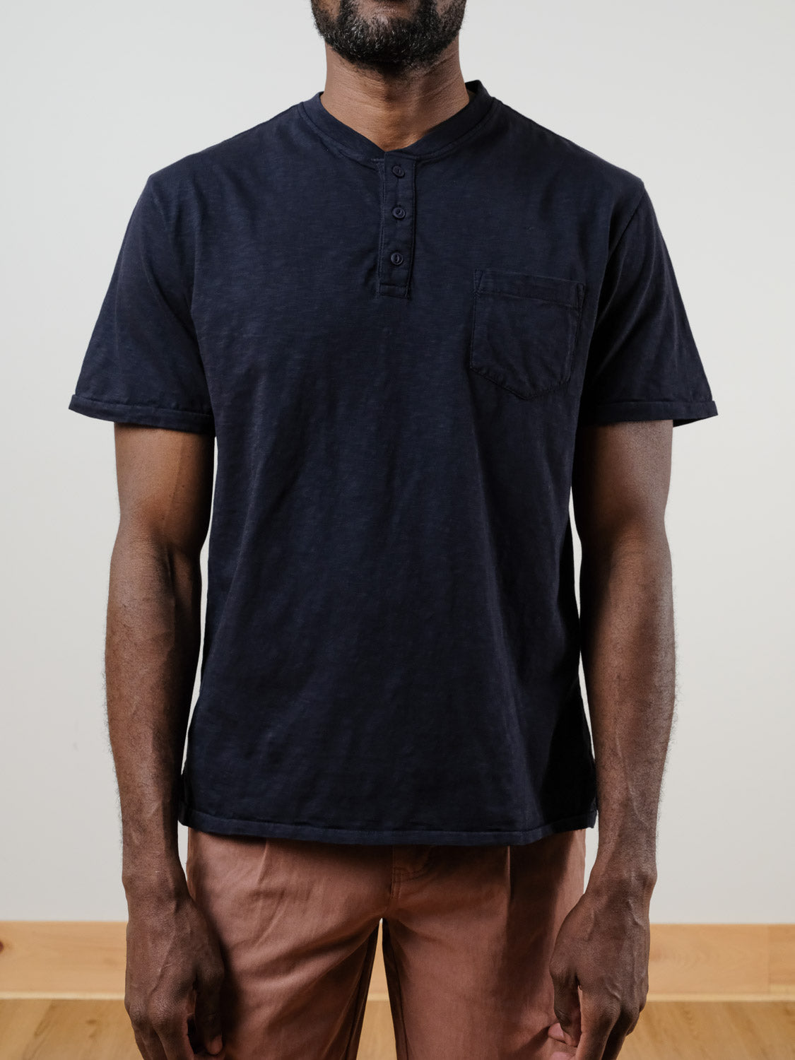 A person, face not visible, wears the Rogue Henley by ID Menswear—an everyday 100% cotton short-sleeve shirt in dark navy with a chest pocket—paired with light brown pants against a plain, light-colored backdrop.