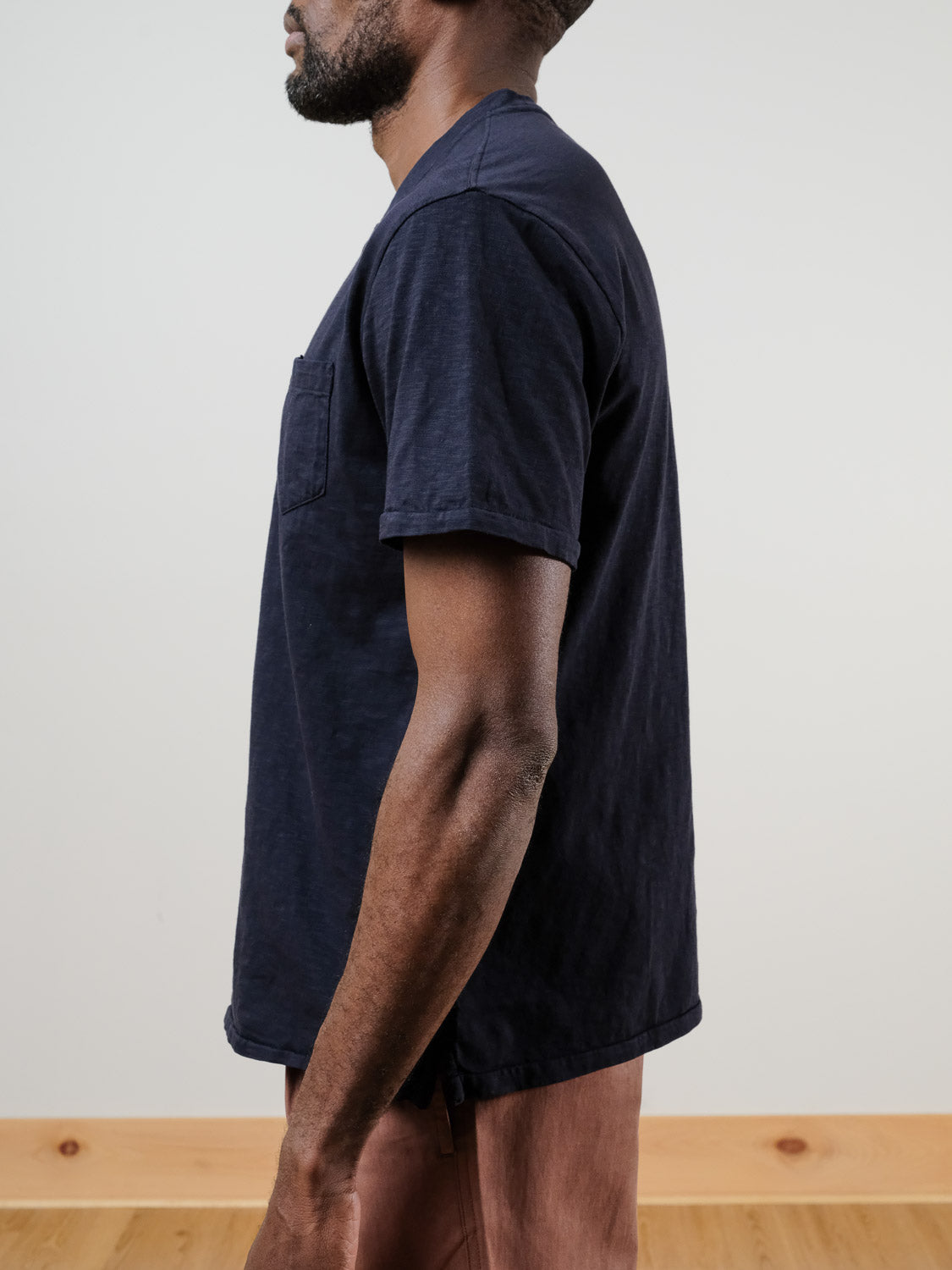 A bearded man wearing the ID Menswear Rogue Henley in navy blue (100% cotton) and brown pants stands in profile, facing left against a plain light background.