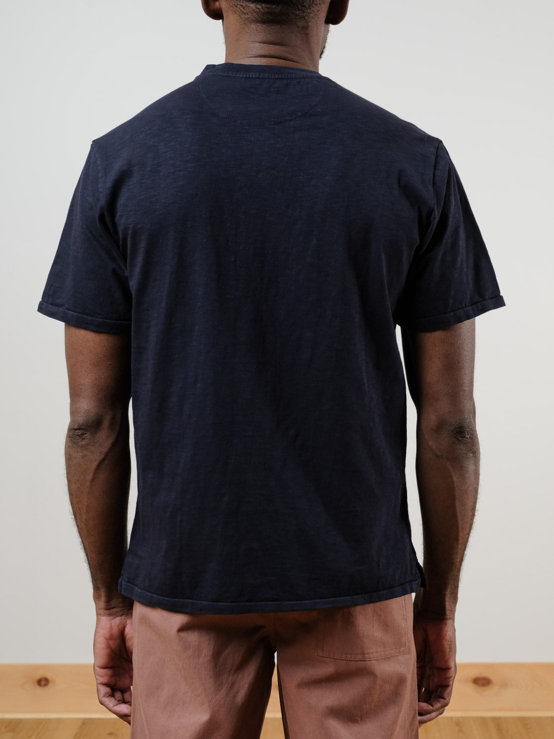 A person stands facing away from the camera, wearing the ID Menswear Rogue Henley in dark navy 100% cotton with light brown pants, set against a plain light background.