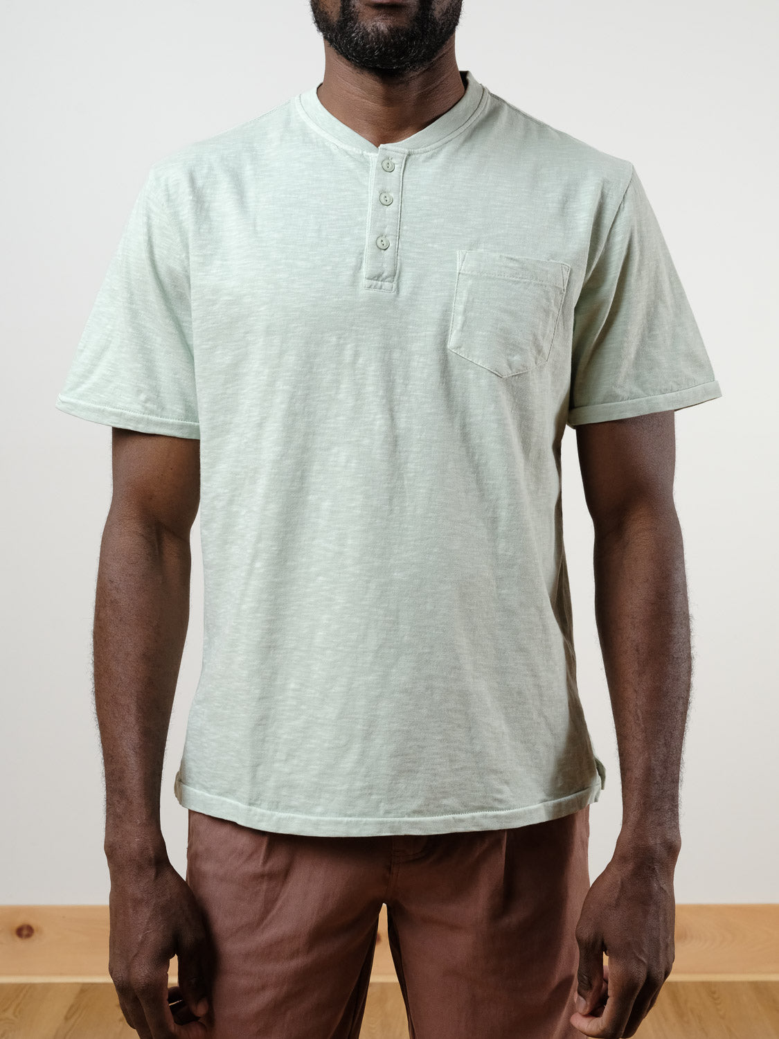 A man stands indoors against a plain light background wearing the ID Menswear Rogue Henley in light green—a short-sleeve, 100% cotton everyday shirt with a chest pocket—paired with brown pants.